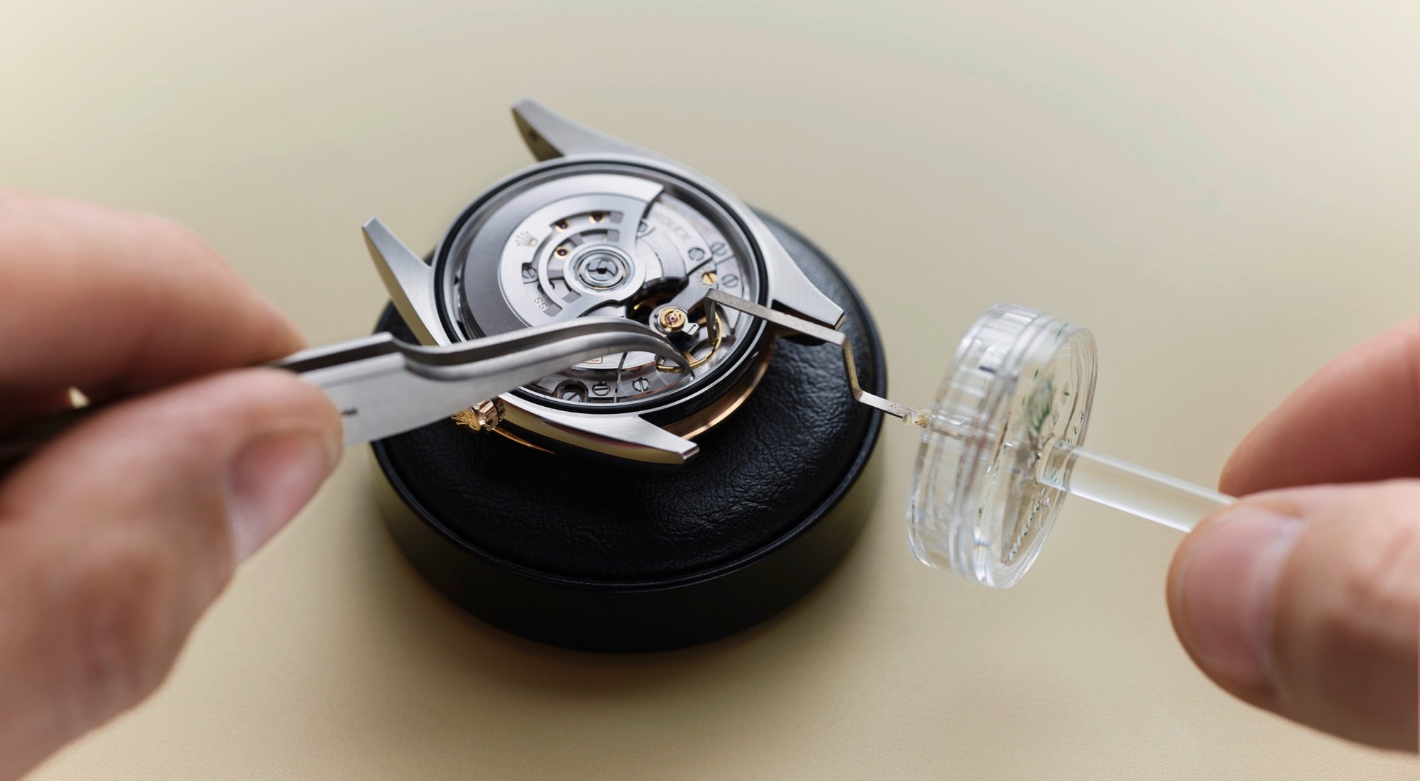 Eine Nahaufnahme eines Uhrmachers, der mit einer Pinzette und einem kleinen Spender vorsichtig Schmiermittel auf die beweglichen Teile eines Uhrwerks aufträgt, das auf einem schwarzen Ständer steht.