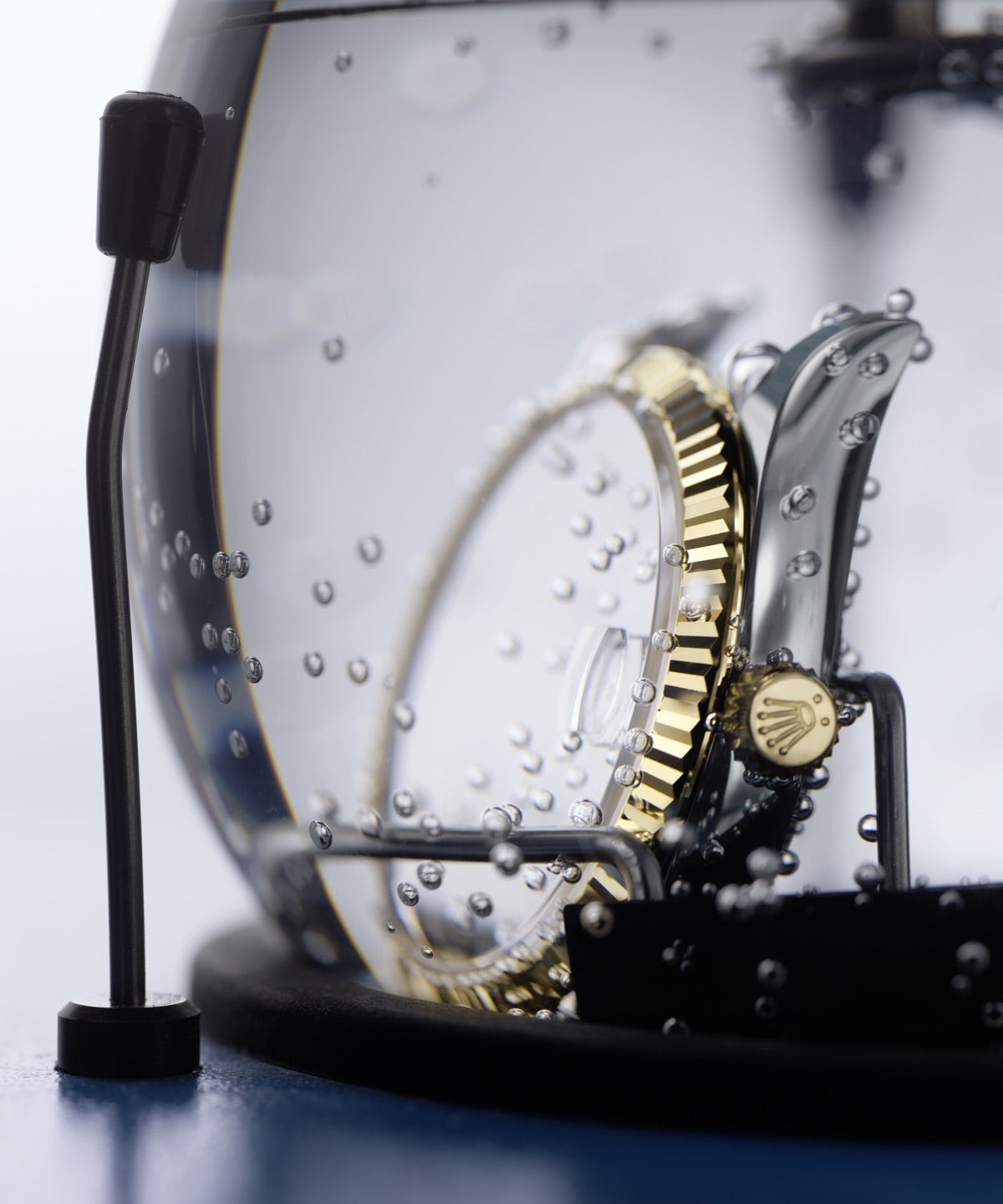 Eine Luxusuhr mit goldener und silberner Lünette wird einem Wasserdichtigkeitstest unterzogen. Sie wird in ein transparentes Becken mit sichtbaren Luftblasen getaucht. Die Krone der Uhr ist mit einem markanten Logo versehen.