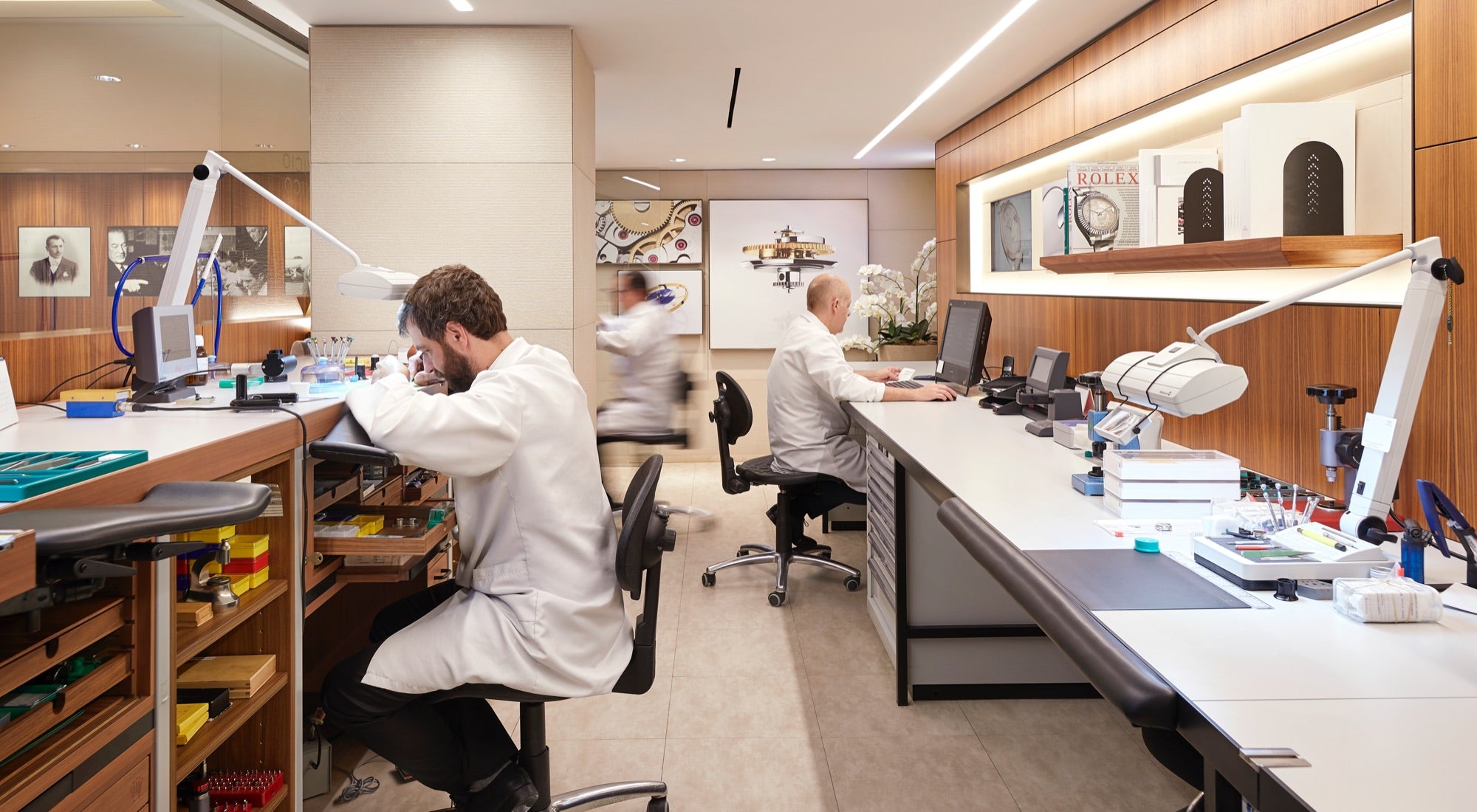 Eine moderne Uhrmacherwerkstatt mit Technikern in weißen Laborkitteln an Schreibtischen. Der Raum ist mit Holzakzenten, Spezialwerkzeugen, Mikroskopen und Regalen mit ordentlich angeordneten Geräten ausgestattet. Eine verschwommene Gestalt geht durch den Raum.