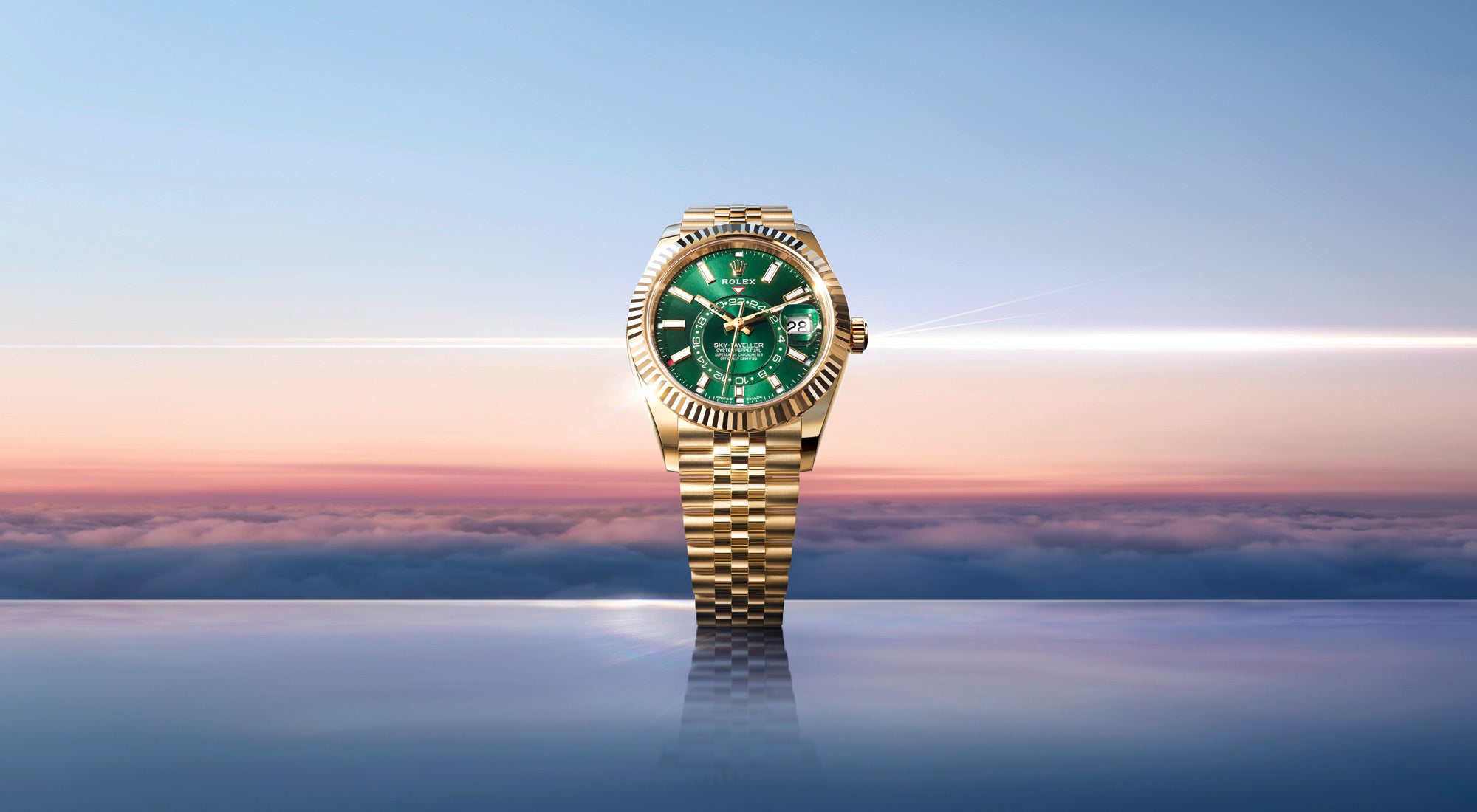 Eine goldene Armbanduhr mit grünem Zifferblatt wird vor einem Hintergrund aus Wolken und einem farbenfrohen Himmel bei Sonnenauf- oder -untergang gezeigt, der sich auf einer glatten Oberfläche darunter spiegelt.