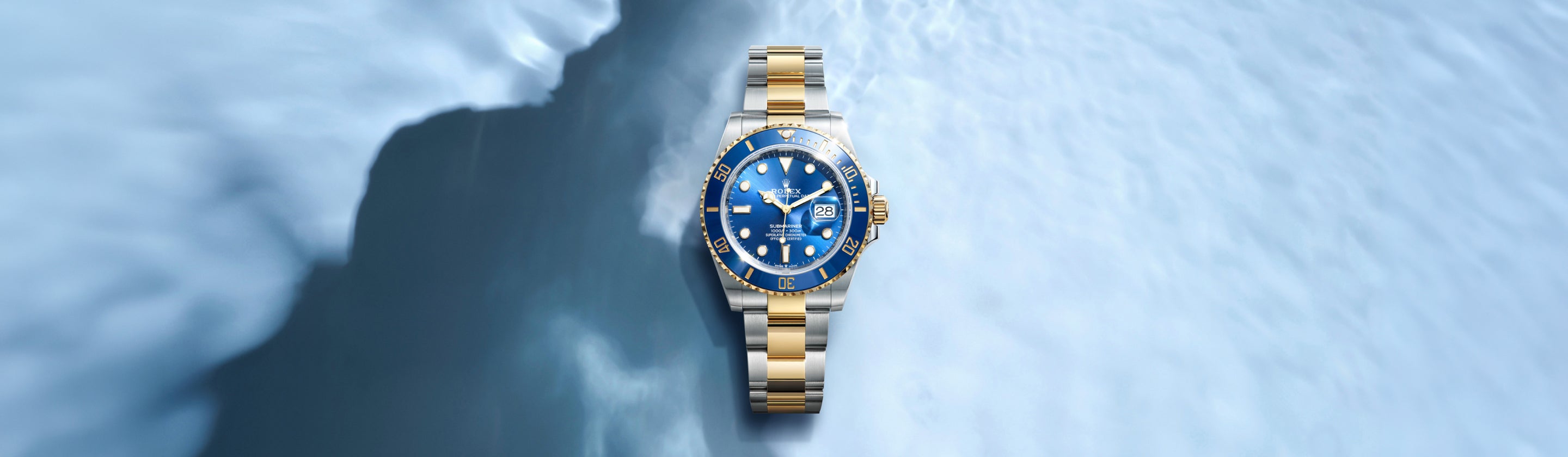 Eine goldene und silberne Armbanduhr mit blauem Zifferblatt und Lünette ruht auf einer blau-weiß strukturierten Oberfläche, die an Eis oder Wasser erinnert. Die Uhr zeigt die Uhrzeit und das Datum an und verfügt über leuchtende Stundenmarkierungen.
