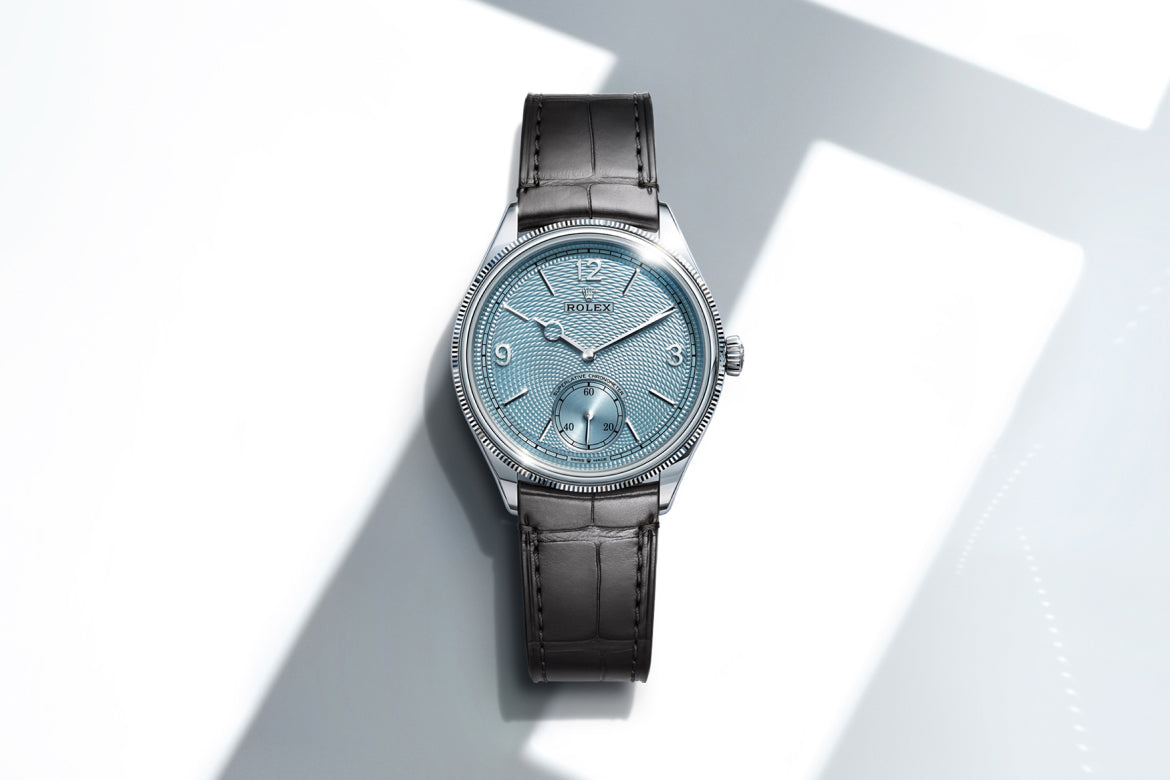 Eine luxuriöse Armbanduhr mit strukturiertem hellblauem Zifferblatt, silbernem Gehäuse und schwarzem Lederarmband wird unter hellem Licht gezeigt und wirft deutliche Schatten auf einen weißen Hintergrund.