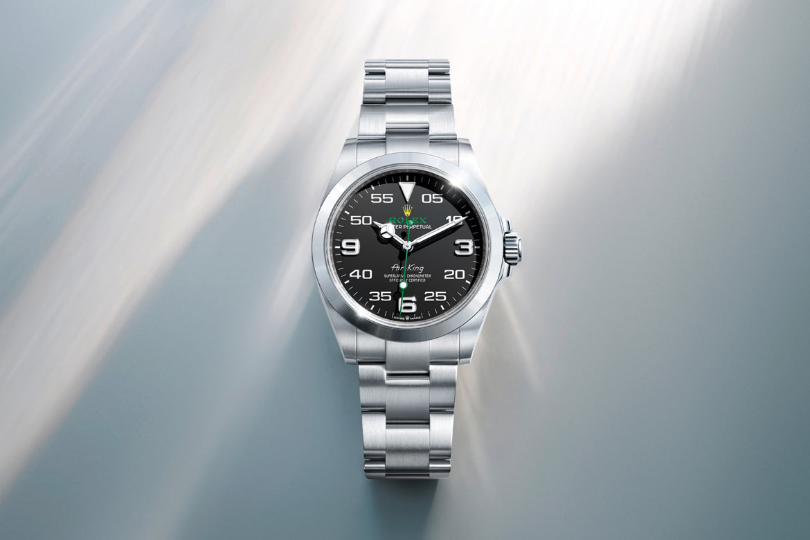 Eine silberne Armbanduhr aus Edelstahl mit schwarzem Zifferblatt, weißen Ziffern und drei Zeigern, die auf einem hellen, leicht verschwommenen Hintergrund mit Lichtschlieren dargestellt sind.