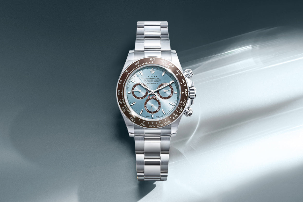 Eine luxuriöse Rolex-Armbanduhr mit silbernem Band, eisblauem Zifferblatt, drei Hilfszifferblättern und einer braunen Keramiklünette, die vor einem sanften blaugrauen Hintergrund mit hellen Streifen steht.