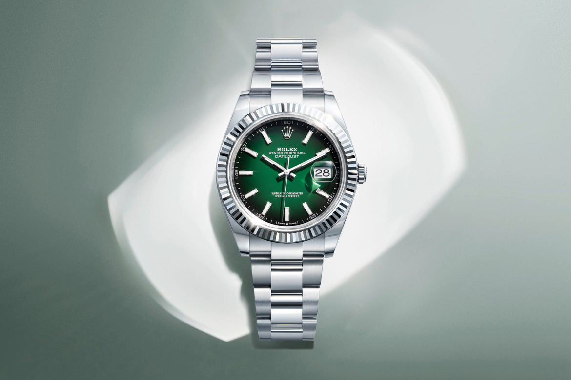 Eine silberne Rolex-Armbanduhr mit Metallband und grünem Zifferblatt wird auf einer hellen, glänzenden Oberfläche mit weichen, weißen Reflexen im Hintergrund dargestellt. Das Zifferblatt ist mit einem Datumsfenster und silbernen Stundenmarkierungen versehen.