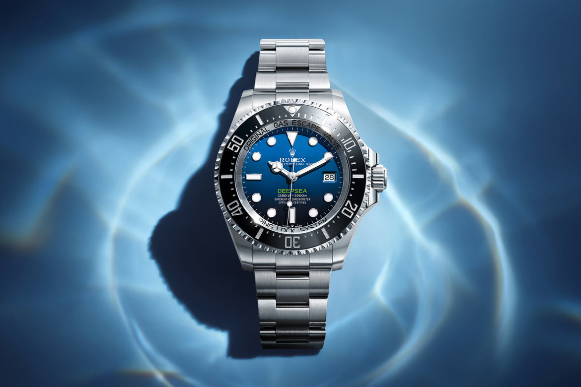 Eine silberne Rolex Deepsea Uhr mit blau-schwarzem Zifferblatt ruht auf einer Oberfläche mit kräuselnden Wasserreflexen. Die Uhr zeigt das Datum an und verfügt über markante Stundenmarkierungen und eine drehbare Lünette.