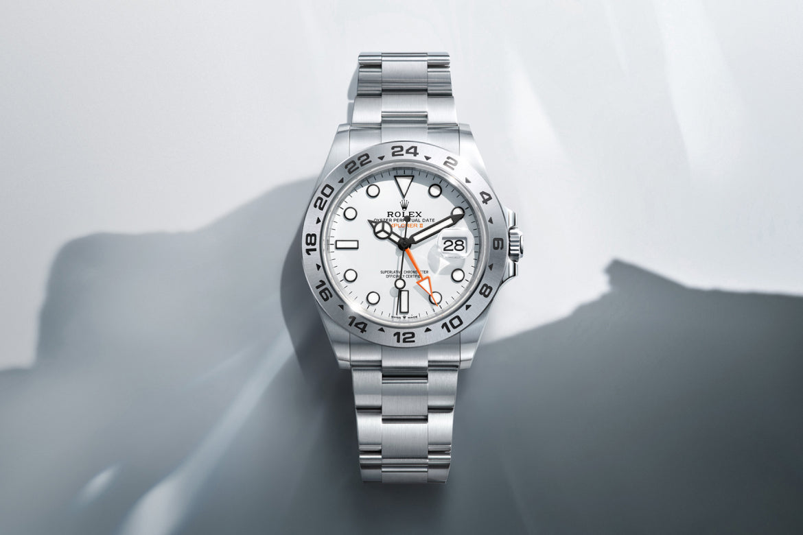 Eine silberne Rolex Explorer II Uhr mit weißem Zifferblatt, schwarzen Stundenmarkierungen und einem orangefarbenen 24-Stunden-Zeiger an einem passenden Metallarmband vor einem hellgrauen, schattigen Hintergrund.