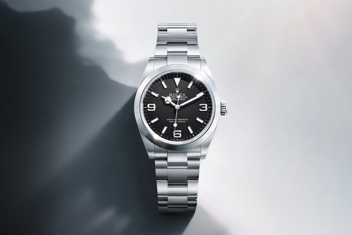 Eine Rolex-Armbanduhr aus Edelstahl mit schwarzem Zifferblatt, silbernen Indexen und einem Metallarmband, dargestellt auf einer glatten Oberfläche mit Schatten und weichem Licht.