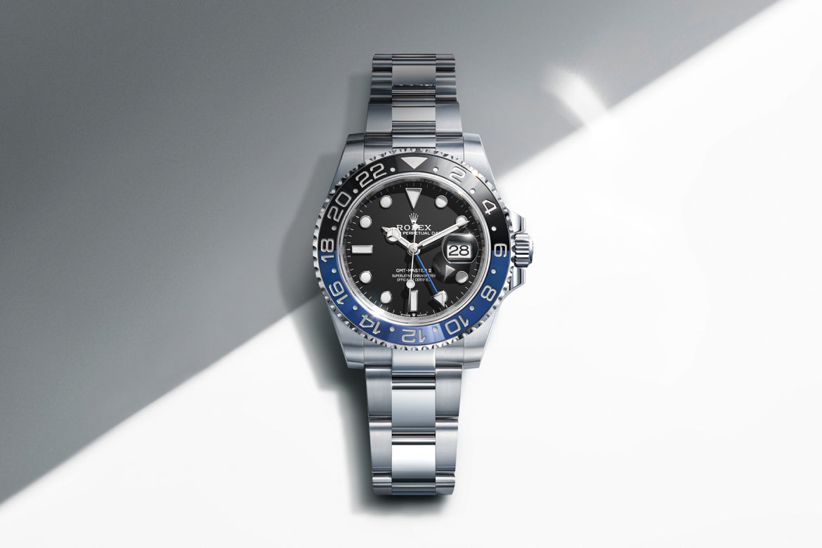 Eine silberne Rolex GMT-Master II Uhr mit schwarzer und blauer Lünette, schwarzem Zifferblatt und Metallarmband wird auf einem weiß-grauen Hintergrund mit diagonaler Beleuchtung dargestellt.