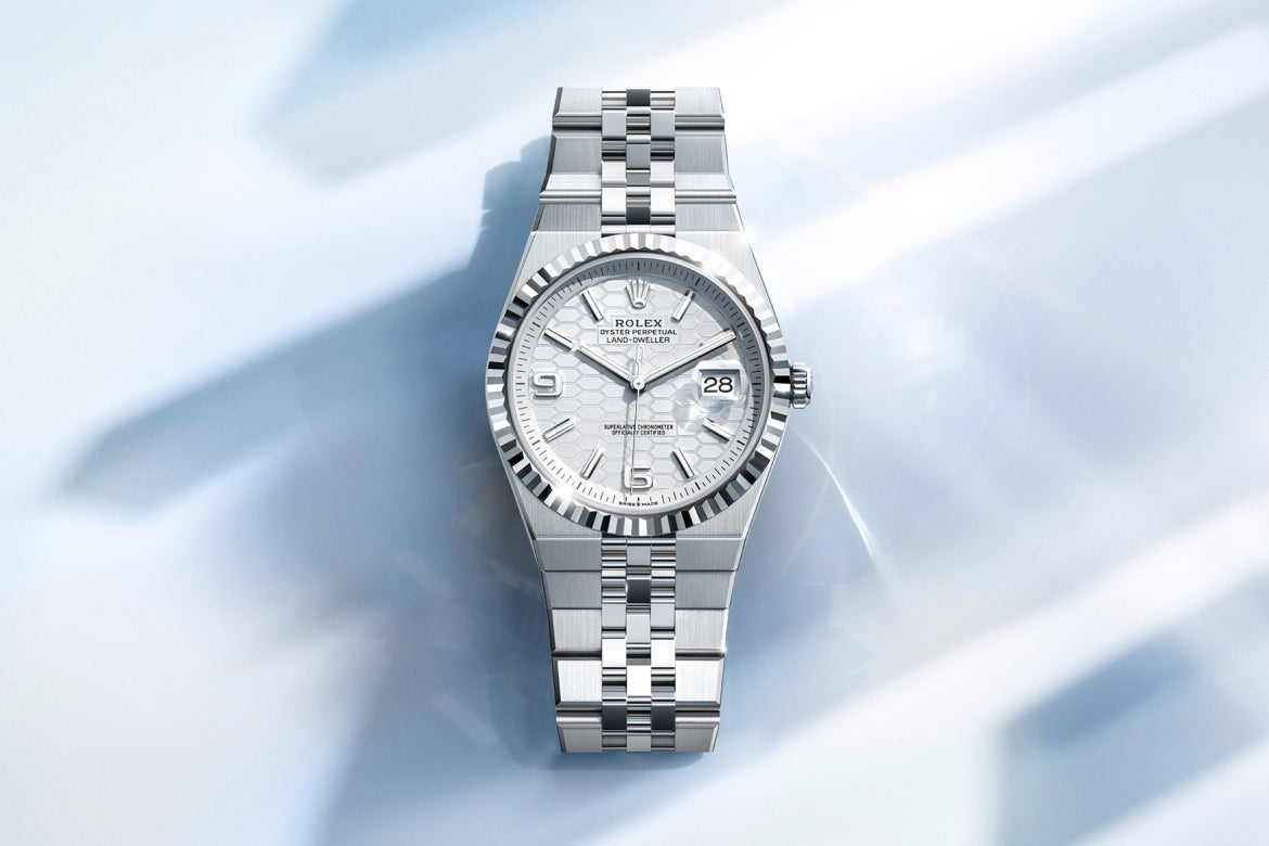 Eine silberne Rolex-Armbanduhr mit Edelstahlband und weißem Zifferblatt wird auf einer hellen, reflektierenden Oberfläche angezeigt. Die Uhr zeigt das Datum an und hat eine geriffelte Lünette mit silbernen Stundenmarkierungen und Zeigern.