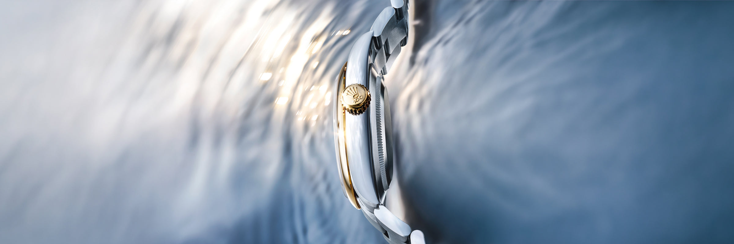 Nahaufnahme einer luxuriösen Armbanduhr mit zweifarbigem Metallband und goldener Krone, in der sich weiches Licht auf einer kräuselnden Wasseroberfläche spiegelt.