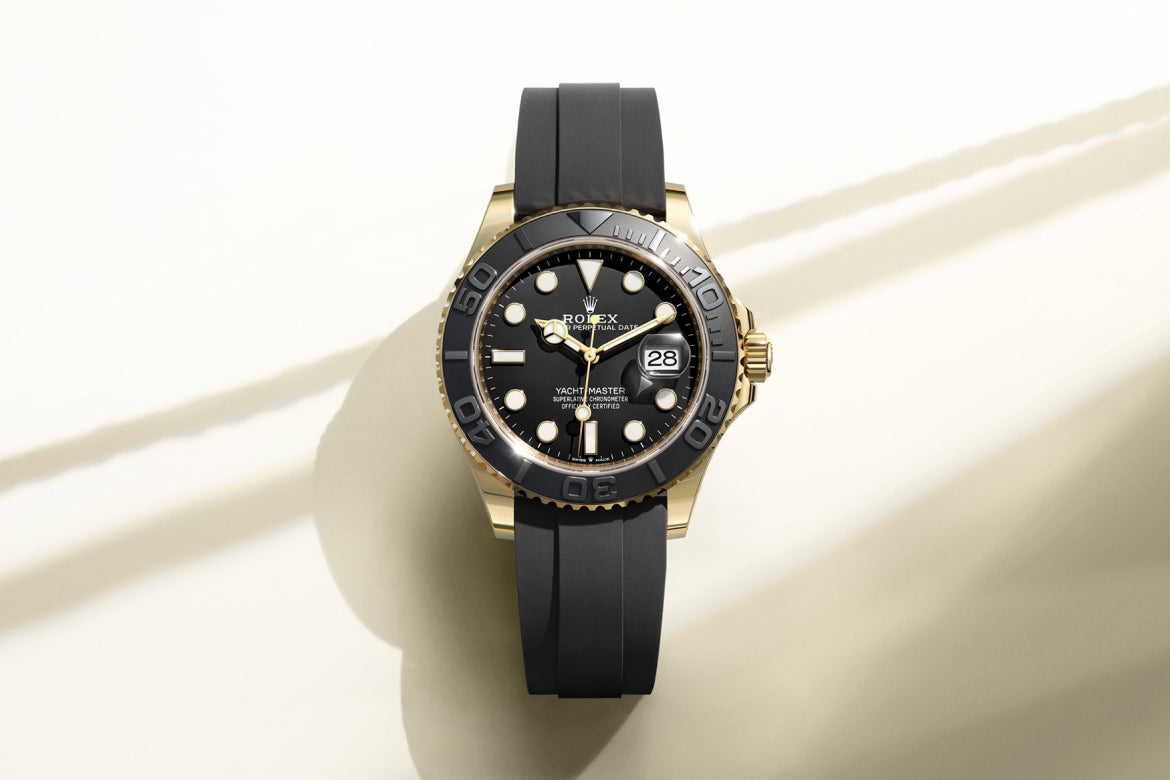 Eine goldene Rolex Yacht-Master Uhr mit schwarzer Lünette, schwarzem Zifferblatt und schwarzem Kautschukarmband wird auf einem hellen Hintergrund mit weichen Schatten dargestellt. Das Datum ist bei der 3-Uhr-Position sichtbar.