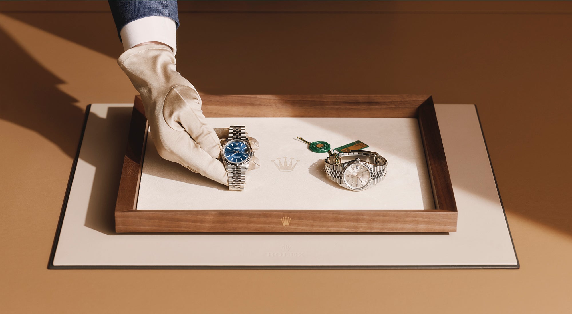 Eine behandschuhte Hand hält eine silberne Uhr mit blauem Ziffernblatt auf einem Tablett, daneben eine weitere silberne Uhr mit weißem Ziffernblatt und grünem Anhänger, die auf einer beigen Matte mit Holzrahmen liegt.