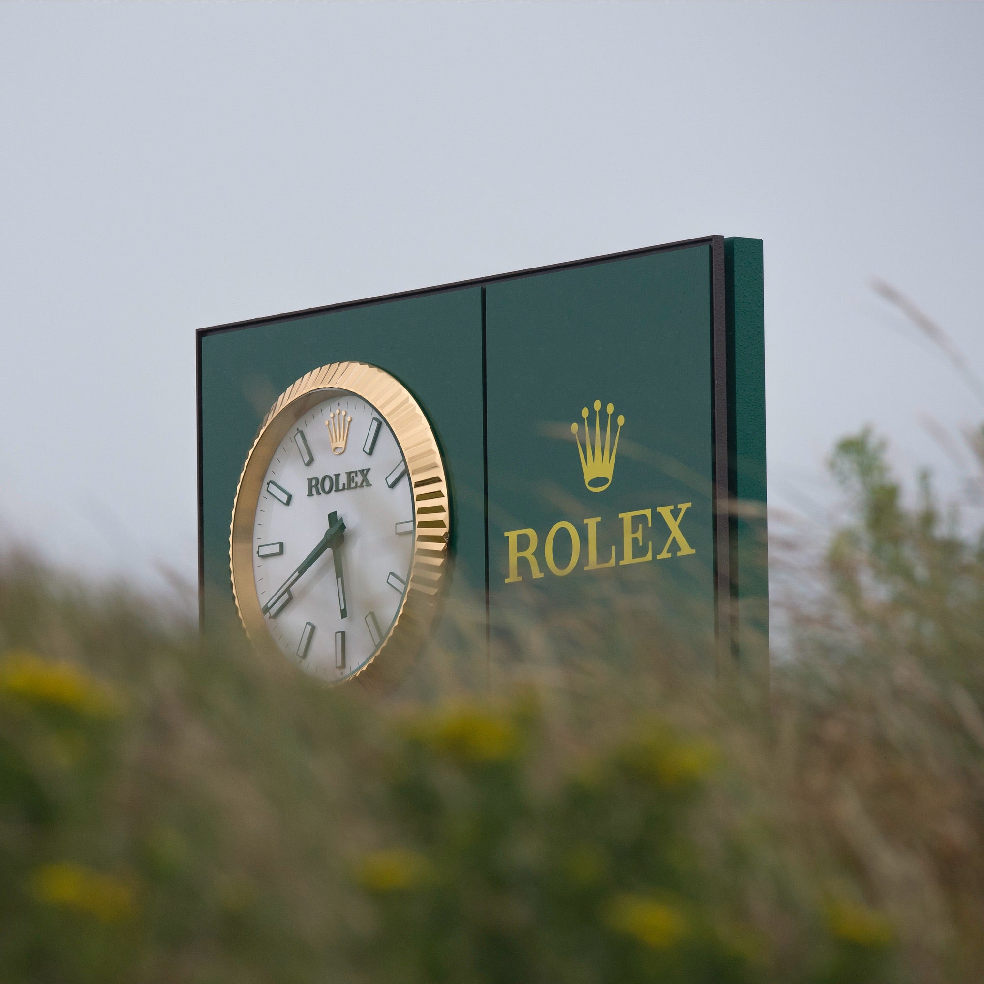 Eine große Rolex-Uhr mit gold-weißem Ziffernblatt ist auf einem grünen Schild mit dem Rolex-Logo und dem Kronenemblem montiert. Der Vordergrund ist mit Grünpflanzen und gelben Blumen verschwommen. Der Himmel ist bedeckt.