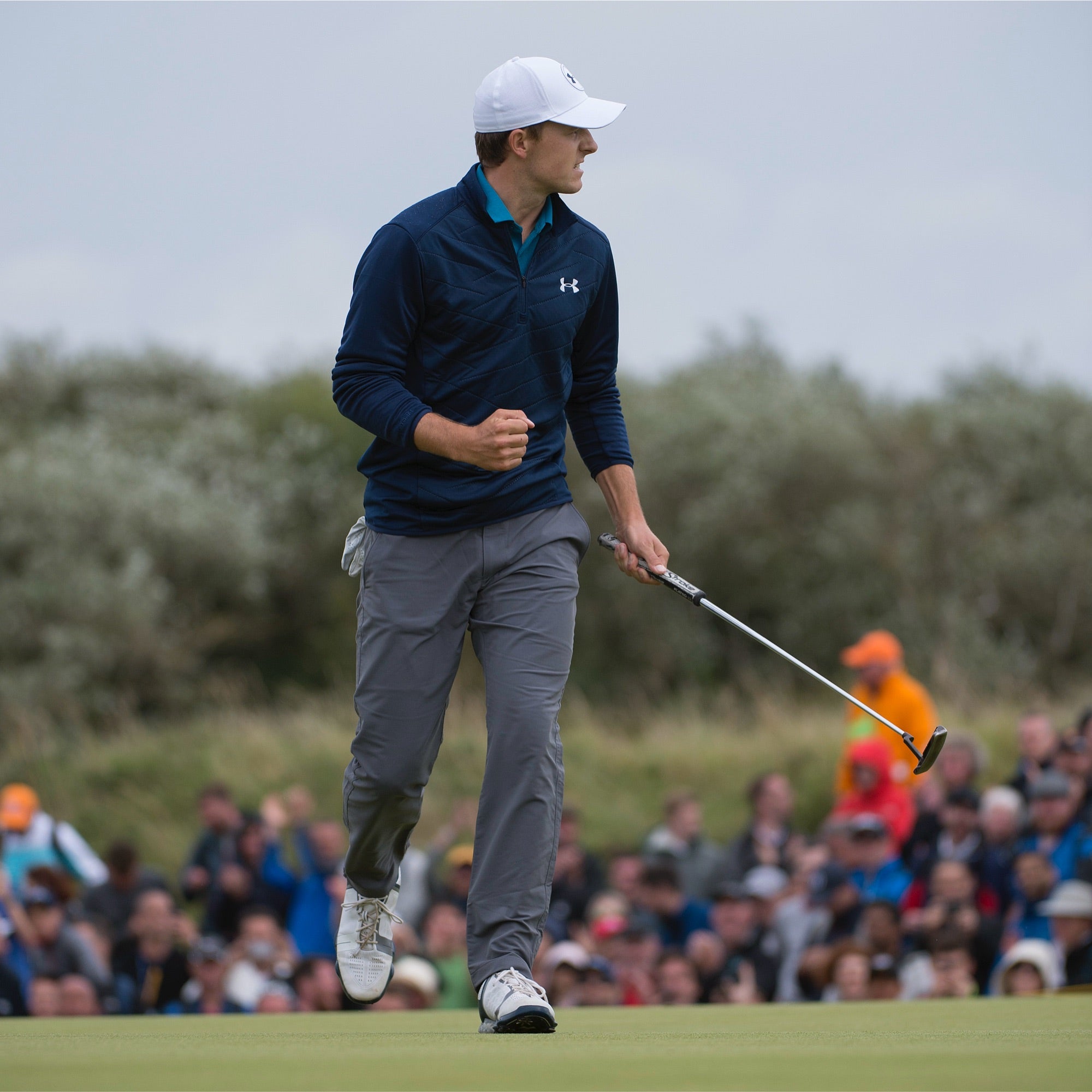 Ein Golfer ballt auf dem Grün jubelnd die Faust und hält einen Putter in der Hand. Er trägt eine marineblaue Jacke, eine weiße Mütze und graue Hosen. Im Hintergrund schaut eine Menschenmenge unter einem bewölkten Himmel zu.