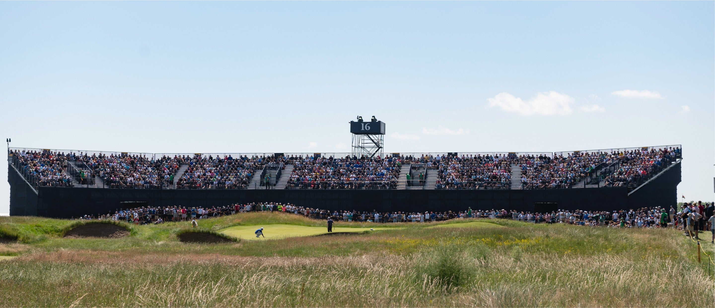 Eine große Menschenmenge in Stadionsitzen verfolgt ein Golfturnier in der Nähe des 16. Lochs. Im Vordergrund ist eine grasbewachsene Golfplatzlandschaft zu sehen. Der Himmel ist klar und blau.
