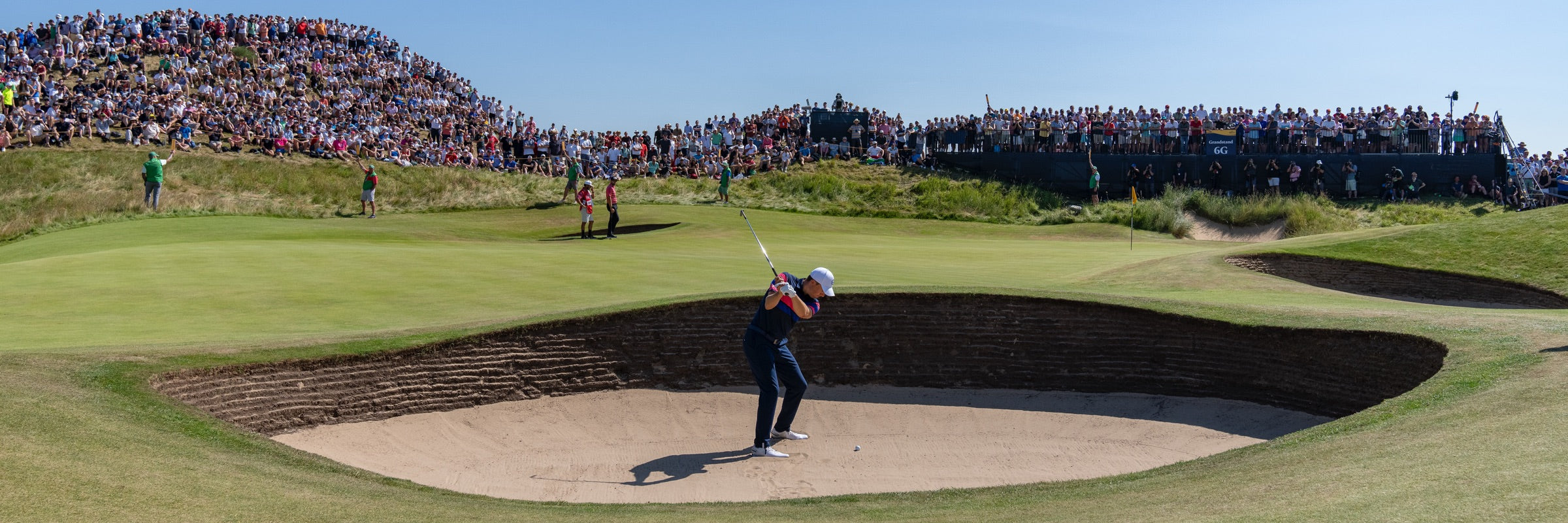 An einem sonnigen Tag auf einem Golfplatz holt ein Golfspieler aus, um einen Schlag aus einem Sandbunker zu machen. Eine große Menge Zuschauer beobachtet ihn vom Rasen aus und steht im Hintergrund.