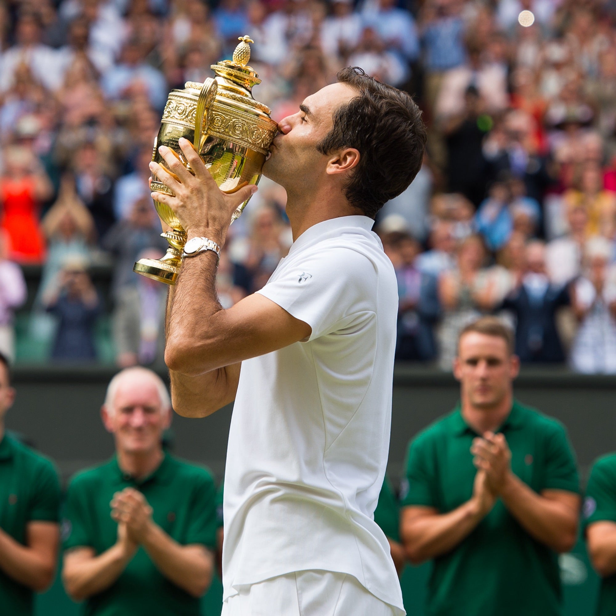Ein Tennisspieler im weißen Outfit küsst auf einem Rasenplatz eine goldene Trophäe. Im Hintergrund applaudieren Zuschauer und Offizielle dem Sieg.