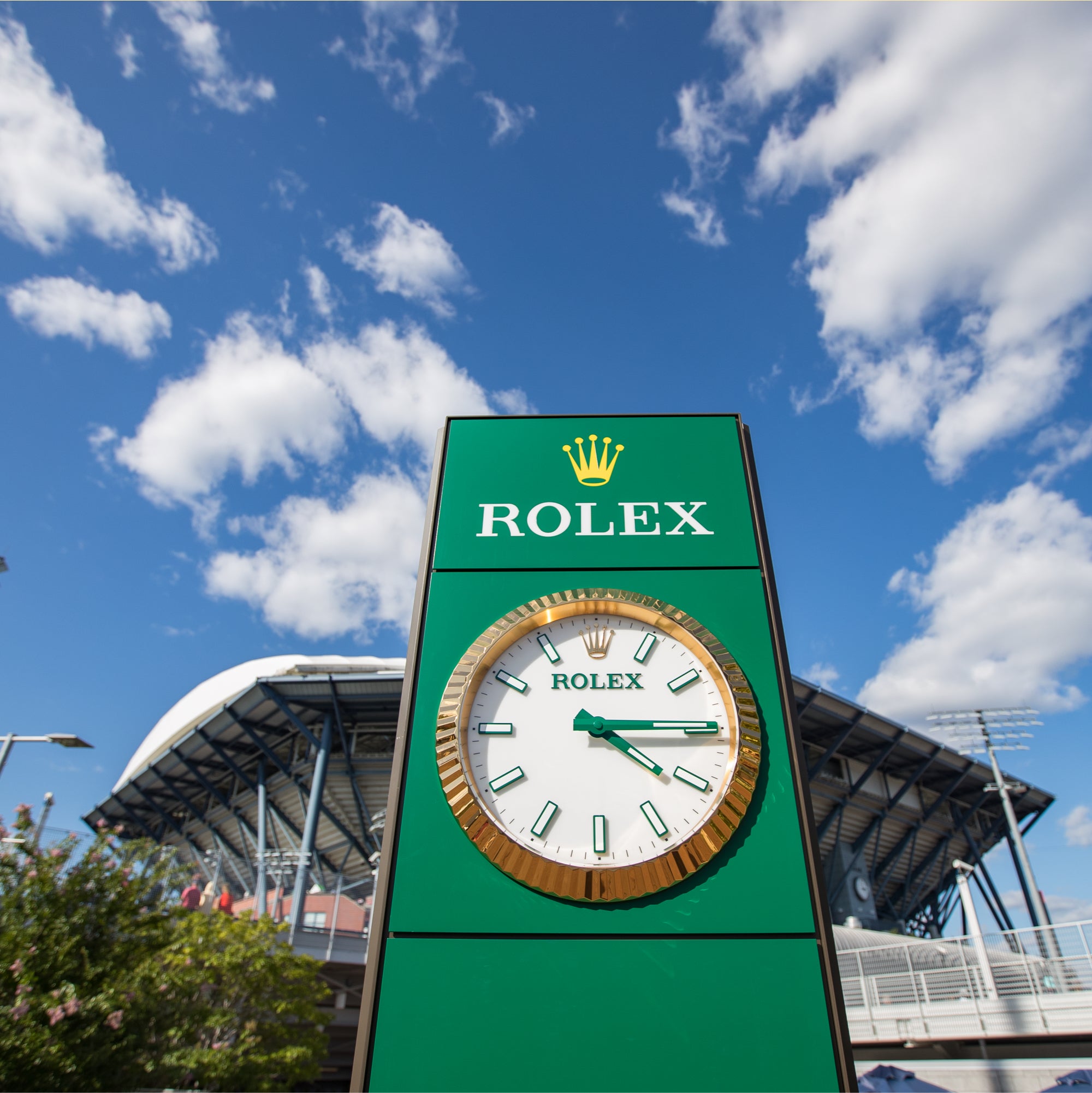 Ein großer grüner Rolex-Uhrenturm steht vor einem blauen Himmel mit vereinzelten Wolken. Im Hintergrund ist ein Stadiongebäude zu erkennen. Die Uhr zeigt ungefähr 1:50 Uhr an.