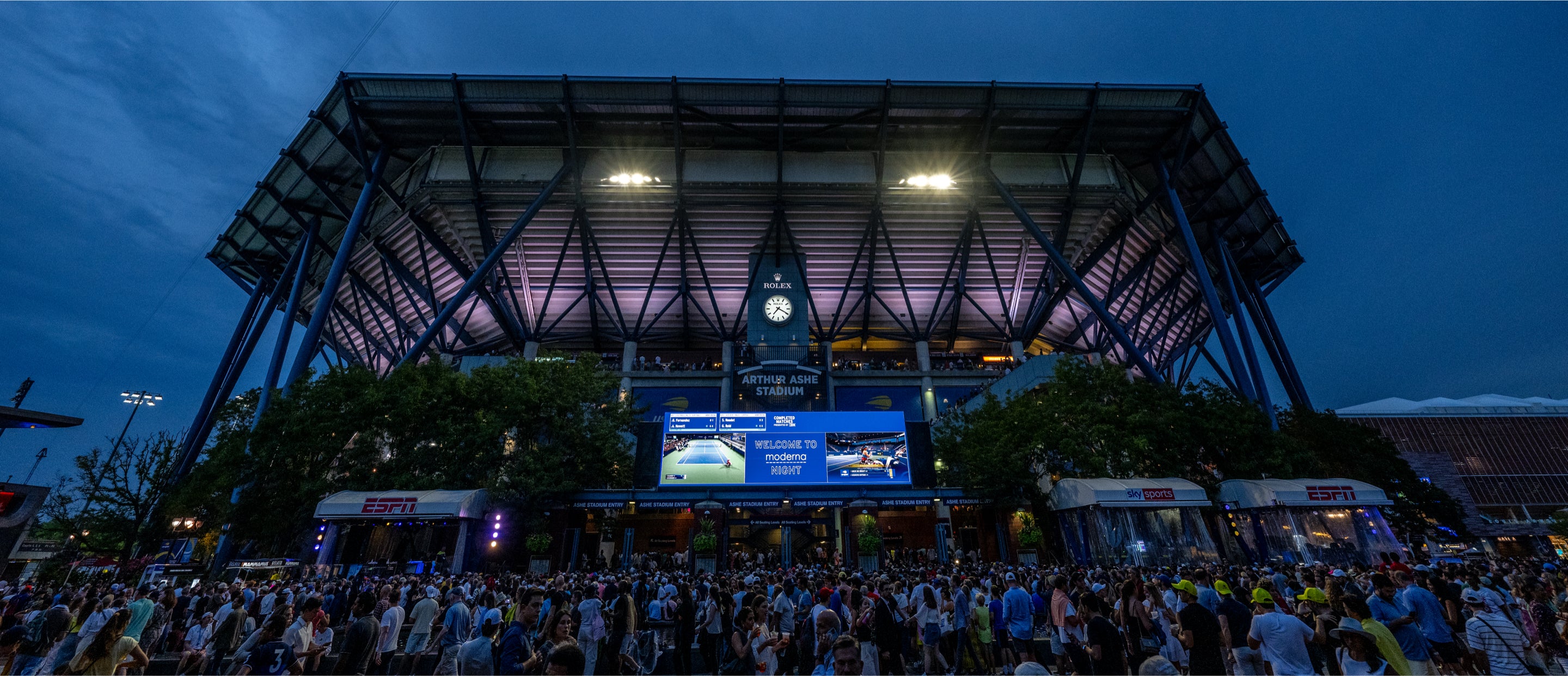 Am Abend versammelt sich eine große Menschenmenge vor dem Arthur Ashe Stadium. Das Stadion ist mit violetten Lichtern beleuchtet und auf digitalen Bildschirmen werden Veranstaltungsinformationen angezeigt. Im Vordergrund sind Bäume und ESPN-Stände zu sehen.