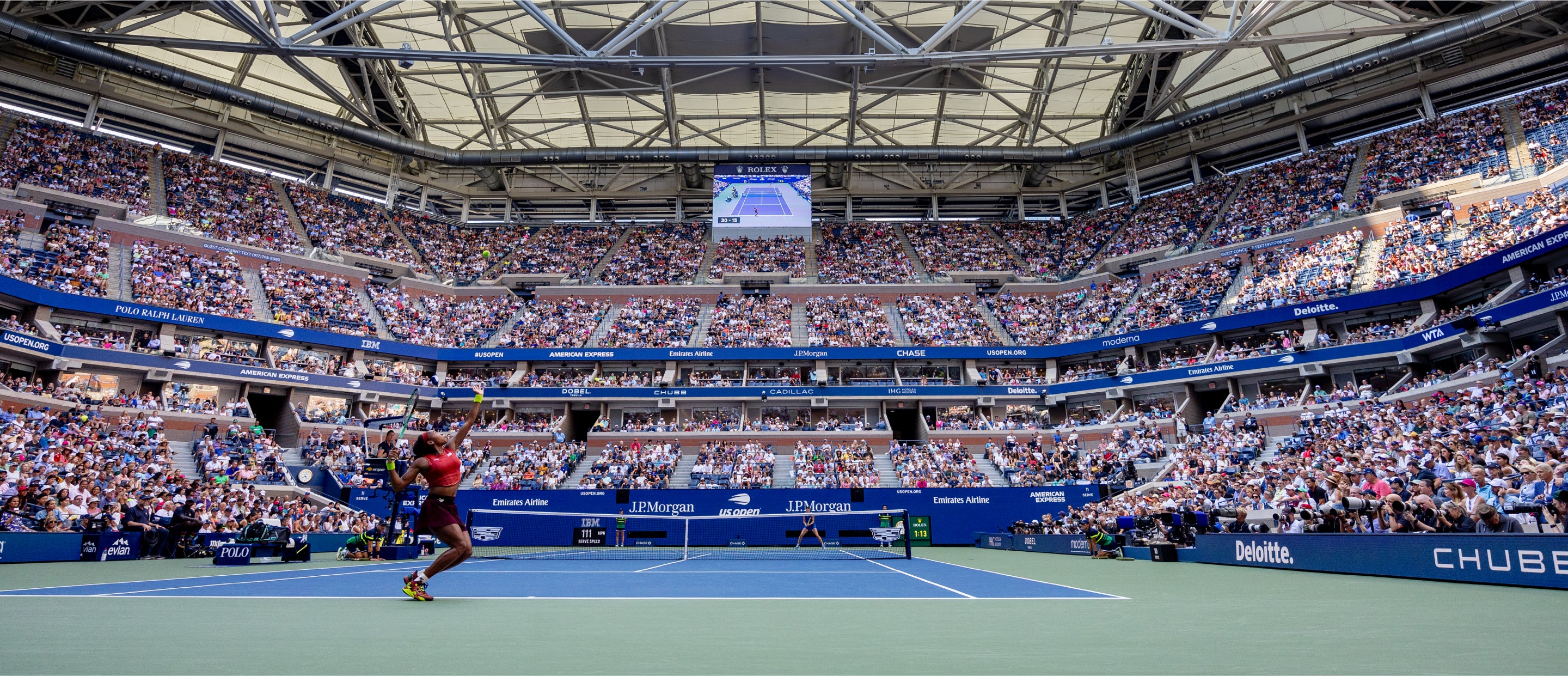 Ein Tennisspieler mitten im Geschehen auf einem blau-grünen Hartplatz in einem großen, voll besetzten Stadion. Die Arena verfügt über mehrere Reihen mit sitzenden Zuschauern und eine Anzeigetafel, auf der die Spieldetails oben angezeigt werden.