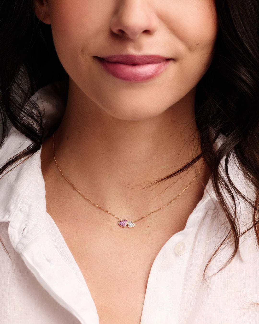 Eine Frau trägt ein weißes Hemd mit Kragen und die Schullin HERZEN HALSKETTE - eine zarte Goldkette mit zwei kleinen herzförmigen Anhängern, einem rosa und einem weißen - und lächelt sanft.