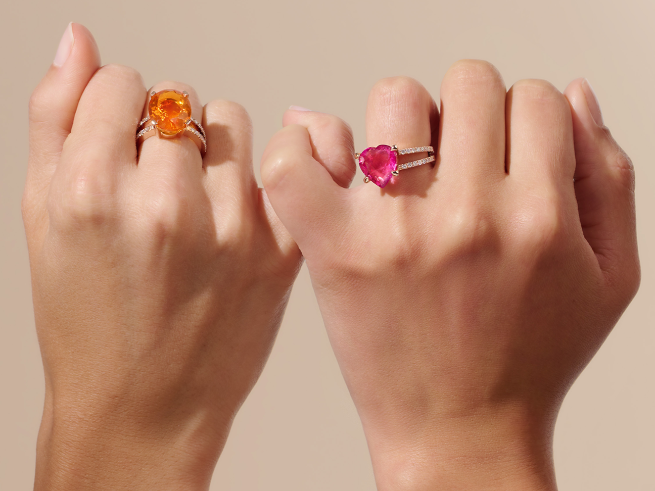 Zwei nach vorne gerichtete Fäuste: Die linke trägt den RUBELLIT RING TWIN von Schullin mit einem großen, runden, orangefarbenen Edelstein, die rechte einen Silberring mit einem rosafarbenen, herzförmigen Edelstein und kleinen, klaren Steinen auf dem Band.