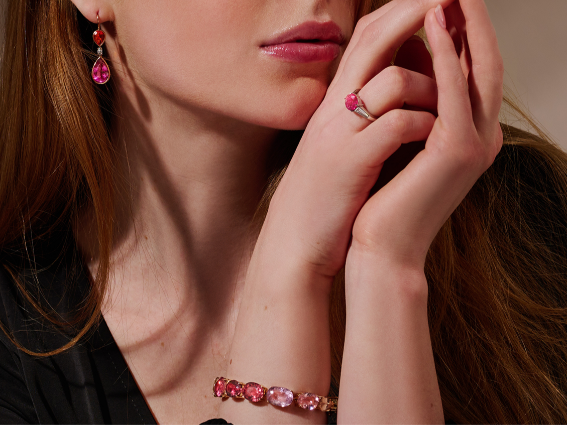 Eine schwarz gekleidete Frau mit langen roten Haaren präsentiert Schullins TURMALIN ARMBAND zusammen mit passenden rosafarbenen Edelsteintropfenohrringen und einem Ring. Sie hält ihre Hände nahe an ihr Gesicht, um die Accessoires hervorzuheben.
