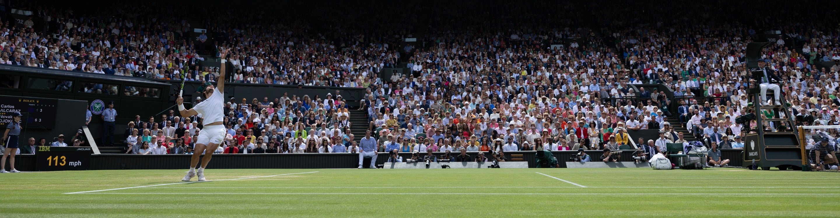 Ein Tennisspieler schlägt in Wimbledon auf einem Rasenplatz auf. Das voll besetzte Stadion schaut gespannt zu, der Schiedsrichter sitzt in der Nähe. Der Spieler ist in vollem Schwung und zeigt Athletik und Konzentration unter hellen Außenbedingungen.