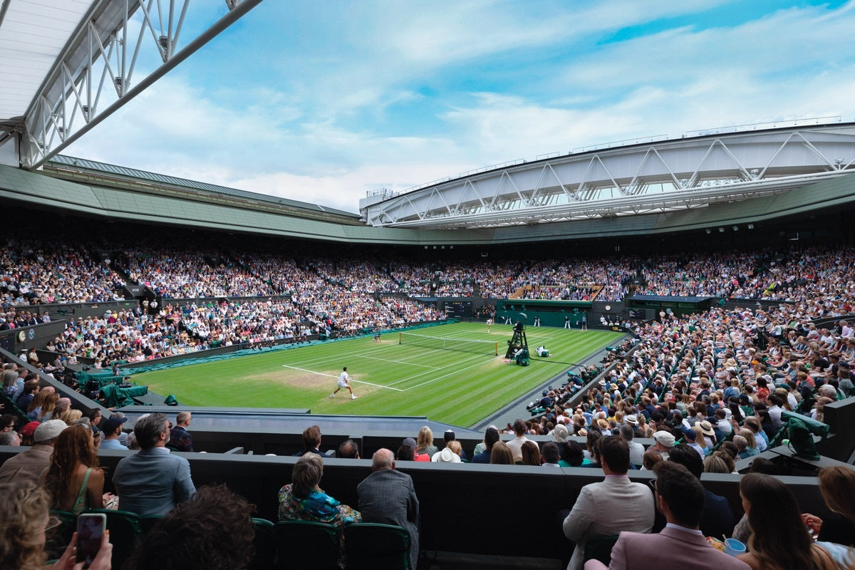 Ein voll besetztes Stadion mit Zuschauern, die auf einem Rasenplatz ein Tennisspiel verfolgen. Der Himmel ist klar und die Arena ist voller Menschen, die auf Sitzreihen sitzen. Die Spieler sind weiß gekleidet und die Atmosphäre ist lebhaft.