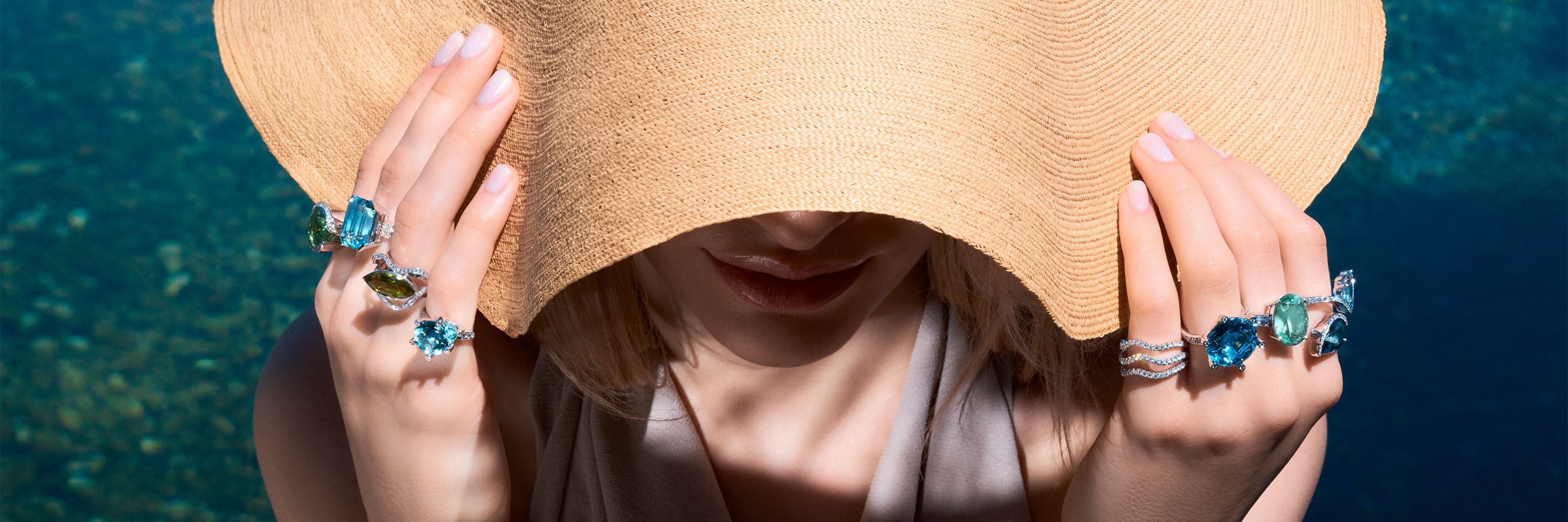 Eine Person mit einem breitkrempigen Hut hält die Krempe mit beiden Händen und präsentiert vor dem Hintergrund des klaren blauen Wassers mehrere große, bunte Edelsteinringe an ihren Fingern.