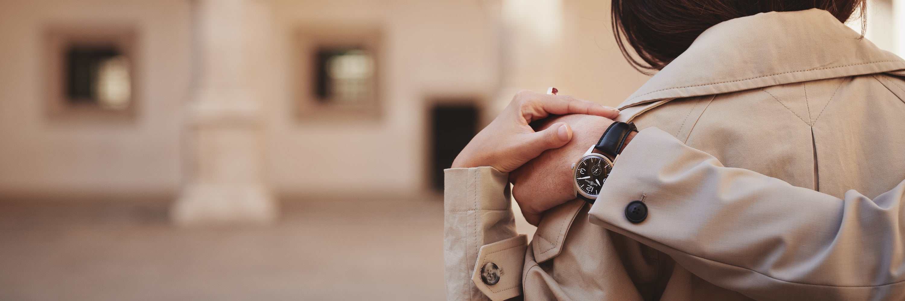 Nahaufnahme einer Person in einem beigen Trenchcoat mit sichtbarer Armbanduhr, die von hinten von einer anderen Person umarmt wird. Der Hintergrund ist verschwommen und zeigt einen Teil eines Gebäudes und einen offenen Innenhof.