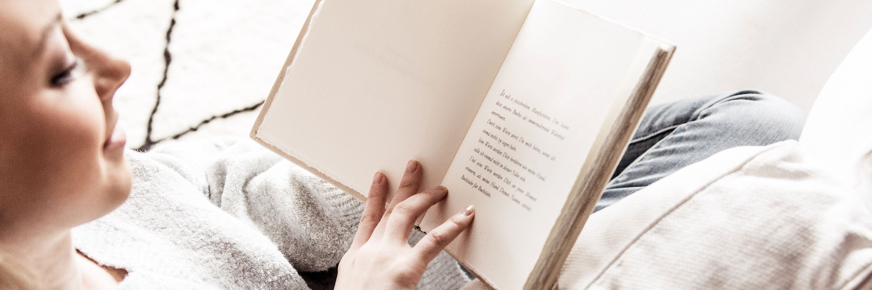 Eine Person liest ein Buch, während sie auf einem Sofa entspannt. Das Buch ist auf einer Seite mit Text aufgeschlagen. Die Person trägt einen weißen Pullover und helle Jeans. Die Atmosphäre wirkt ruhig und gemütlich.