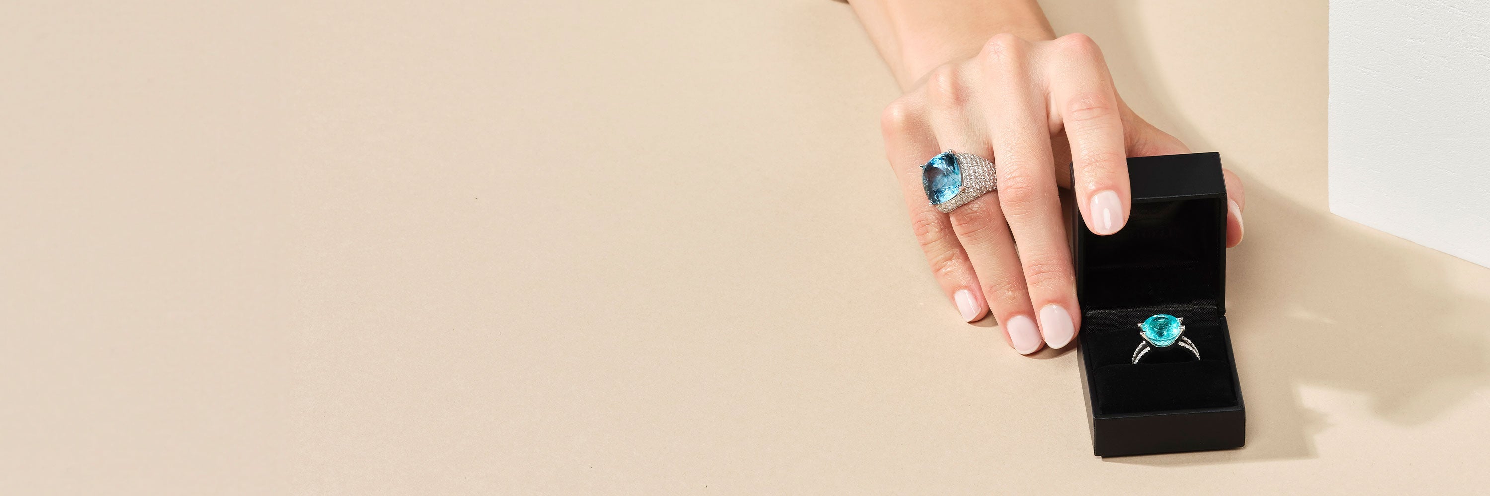 Hinter einer weißen Blende ist teilweise eine Person in einem weißen Langarmhemd zu sehen. An ihren Fingern trägt sie einen Ring mit blauem Stein. In der Hand hält sie eine schwarze Schmuckschatulle mit einem weiteren Ring mit blauen Steinen darin.