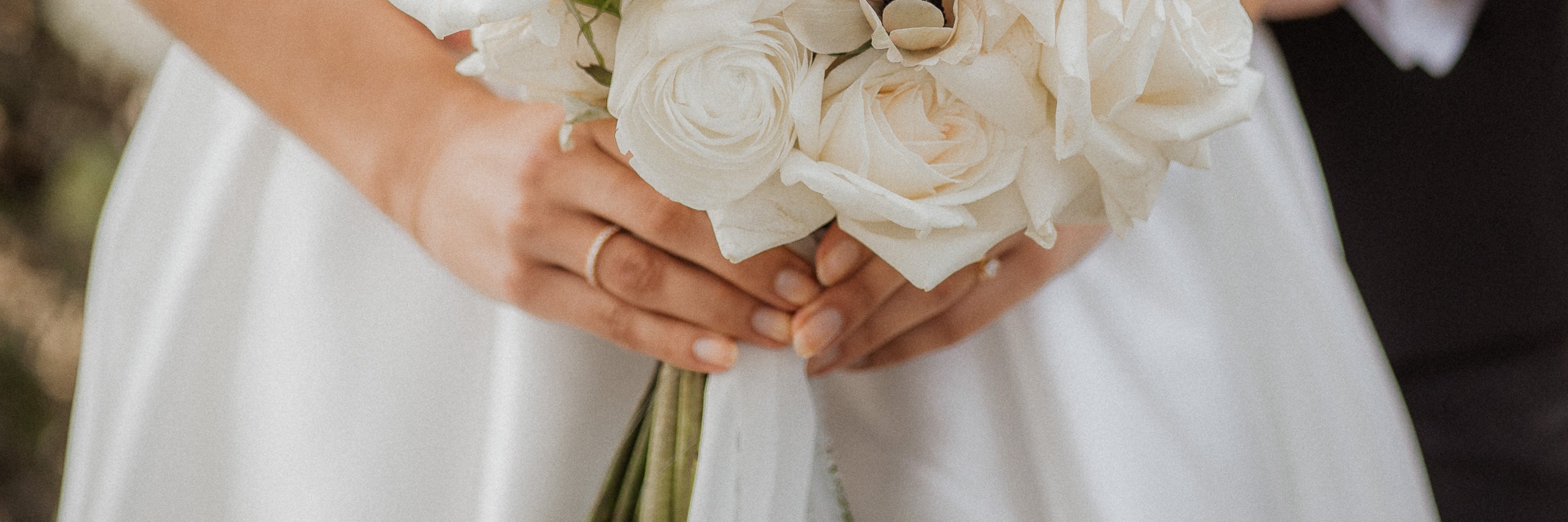 Eine Nahaufnahme einer Braut, die einen Strauß weißer Rosen in beiden Händen hält. Im Hintergrund ist teilweise der Arm des Bräutigams zu sehen. Die Braut trägt ein weißes Kleid und einen schlichten goldenen Ehering.