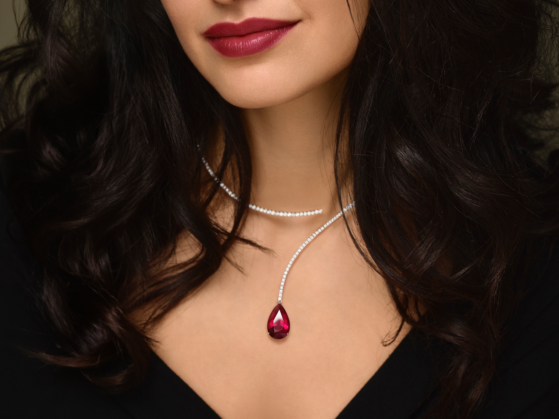 Eine Frau mit gewelltem dunklem Haar trägt tiefroten Lippenstift und das Schullin DIAMANT HALSREIF in Weißgold mit einem großen Rubellit Tropfen-Anhänger. Sie kombiniert die Halskette mit einem schwarzen Oberteil.