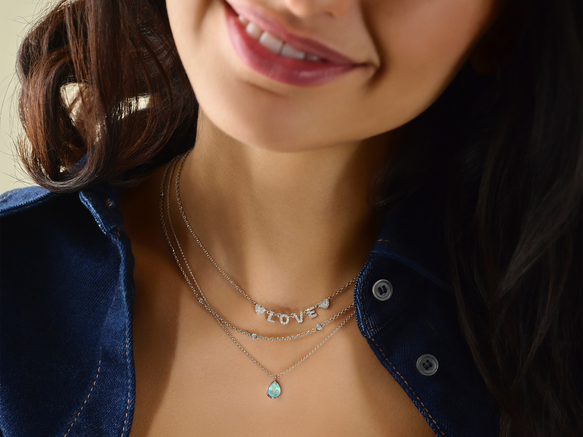 Eine Frau lächelt und trägt die Schullin LOVE HALSKETTE - eine mehrschichtige Silberkette mit Buchstabenanhängern, die "LOVE" bedeuten - zusammen mit einer anderen Kette mit einem blauen Tropfenanhänger und einem blauen, teilweise aufgeknöpften Oberteil.