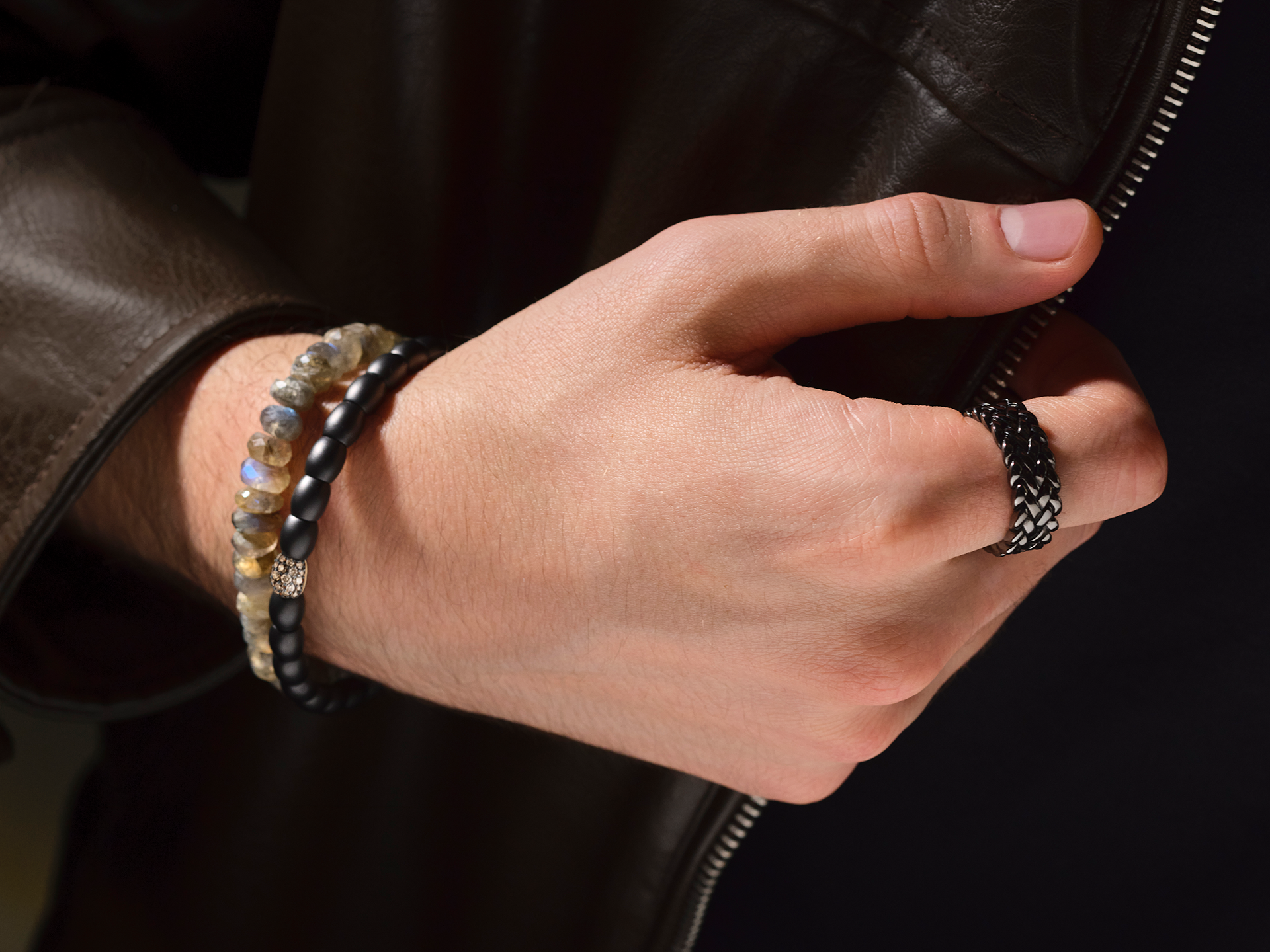 Nahaufnahme einer Hand, die Schullins INTUITION ARMBAND und einen strukturierten Ring trägt und nach dem Reißverschluss einer braunen Lederjacke greift.