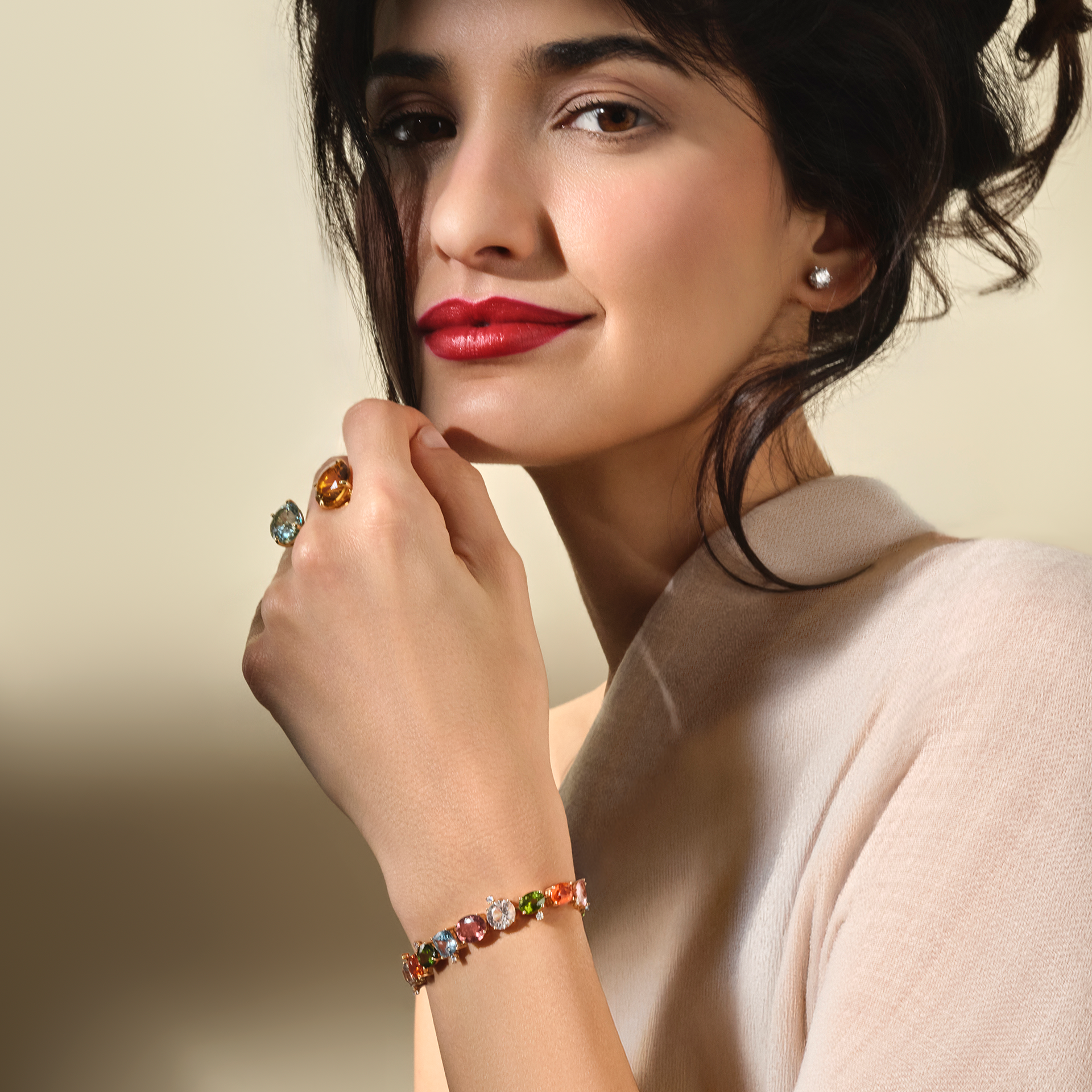 Eine Frau mit rotem Lippenstift und dunklem Haar trägt ein beiges Oberteil, ein Schullin ARMBAND COLOURSPLASH, einen Perlenohrring und einen großen Edelsteinring und lächelt sanft vor einem neutralen Hintergrund.