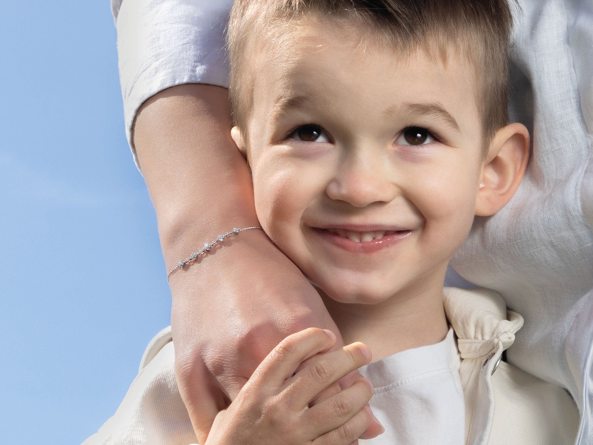Ein kleiner lächelnder Junge hält die Hand eines Erwachsenen und blickt glücklich nach oben. Der Erwachsene trägt ein Schullin MUTTERTAGS SPECIAL ARMBAND an seinem weißärmeligen Handgelenk. Ein klarer blauer Himmel im Hintergrund deutet auf einen sonnigen Tag hin.