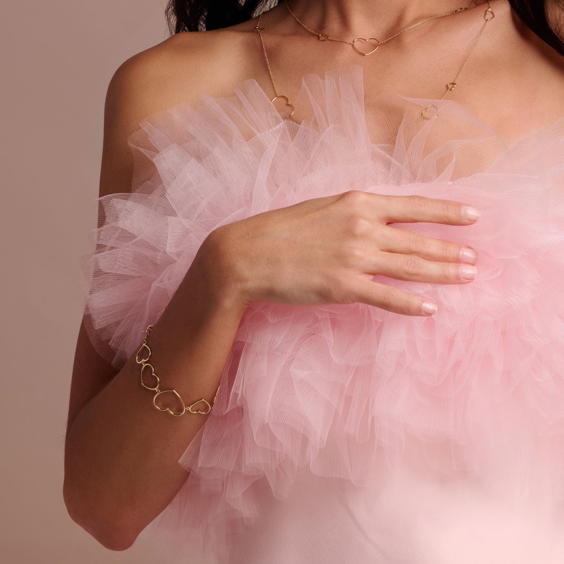 Eine Frau in einem hellrosafarbenen Tüllkleid trägt das goldene HERZEN ARMBAND von Schullin und eine passende herzförmige Halskette. Sie berührt ihre Brust vor einem weichen, neutralrosa Hintergrund.