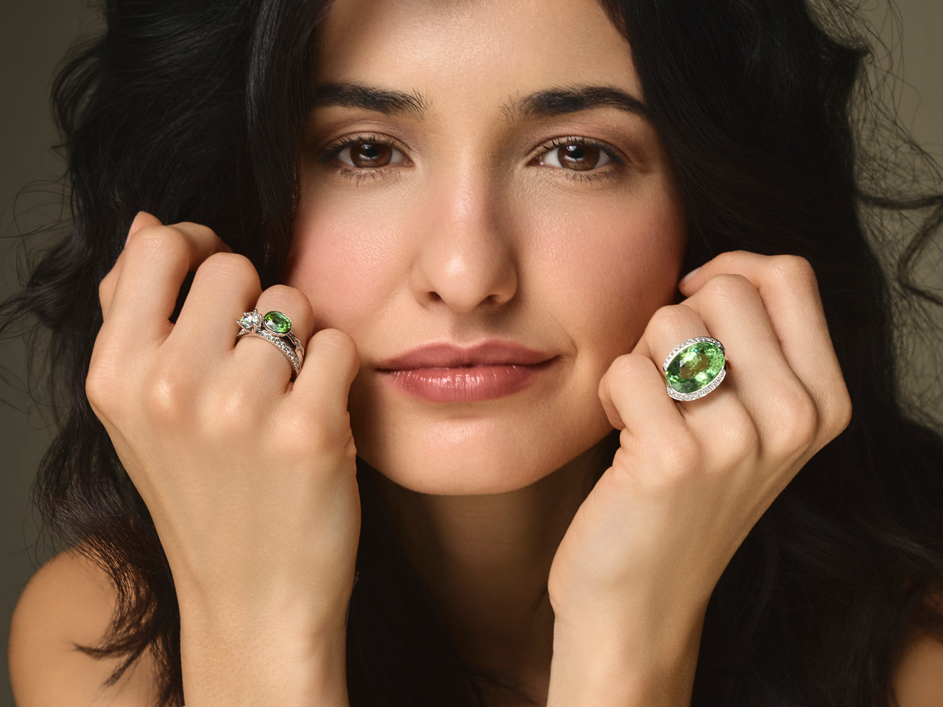 Eine dunkelhaarige Frau hält die Fäuste unter dem Kinn und präsentiert einen Diamantenring und den TURMALIN RING von Schullin mit großen hellgrünen Edelsteinen. Ihr Gesichtsausdruck ist neutral und sie trägt ein sanftes Make-up.