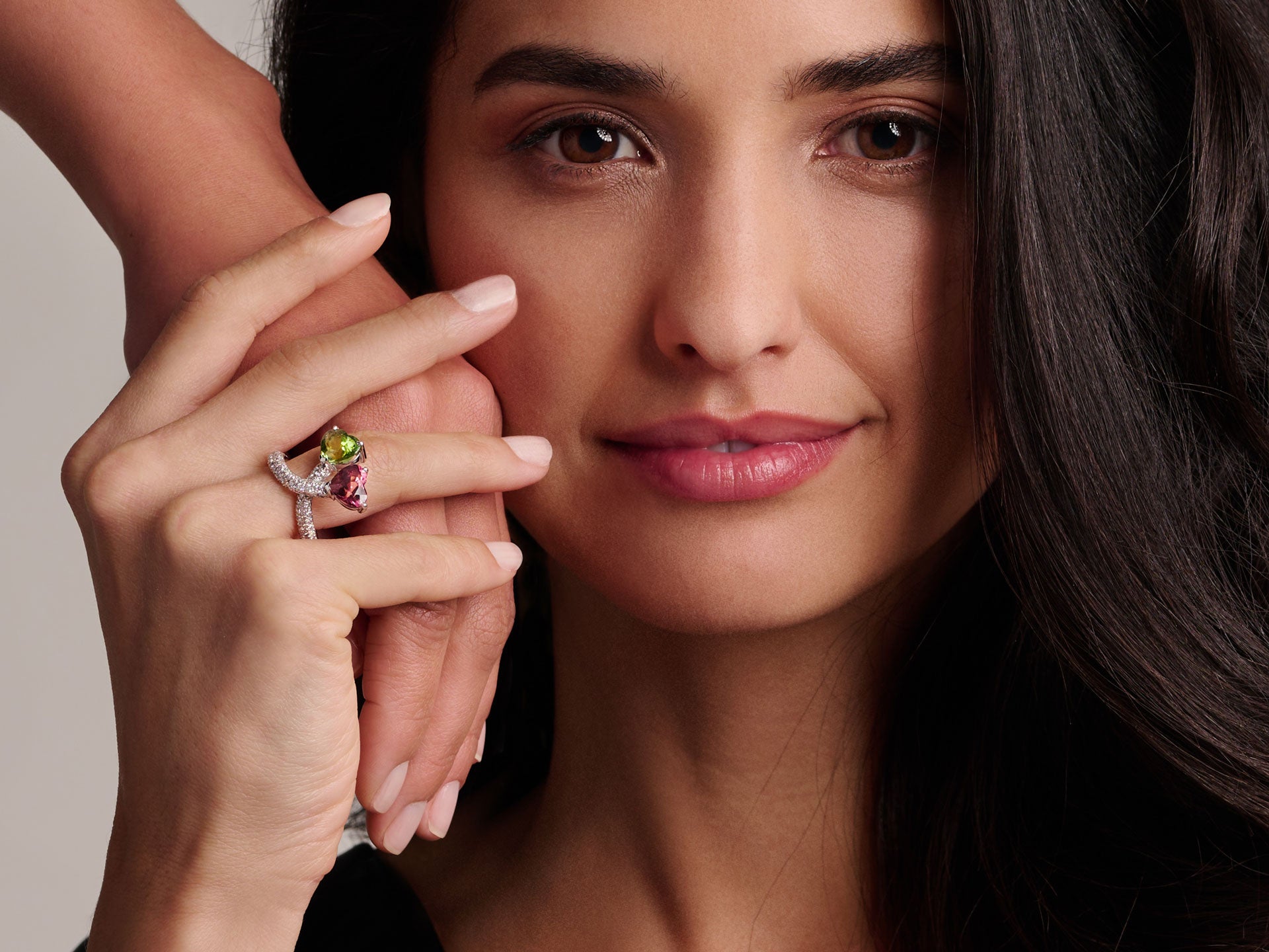 Eine Frau mit langen dunklen Haaren lächelt sanft, hält ihre Hand an ihr Gesicht und trägt den RING TOGETHER von Schullin, einen funkelnden Ring mit grünen und roten Steinen. Eine andere Hand ruht sanft auf ihrem Handgelenk vor einem neutralen Hintergrund.