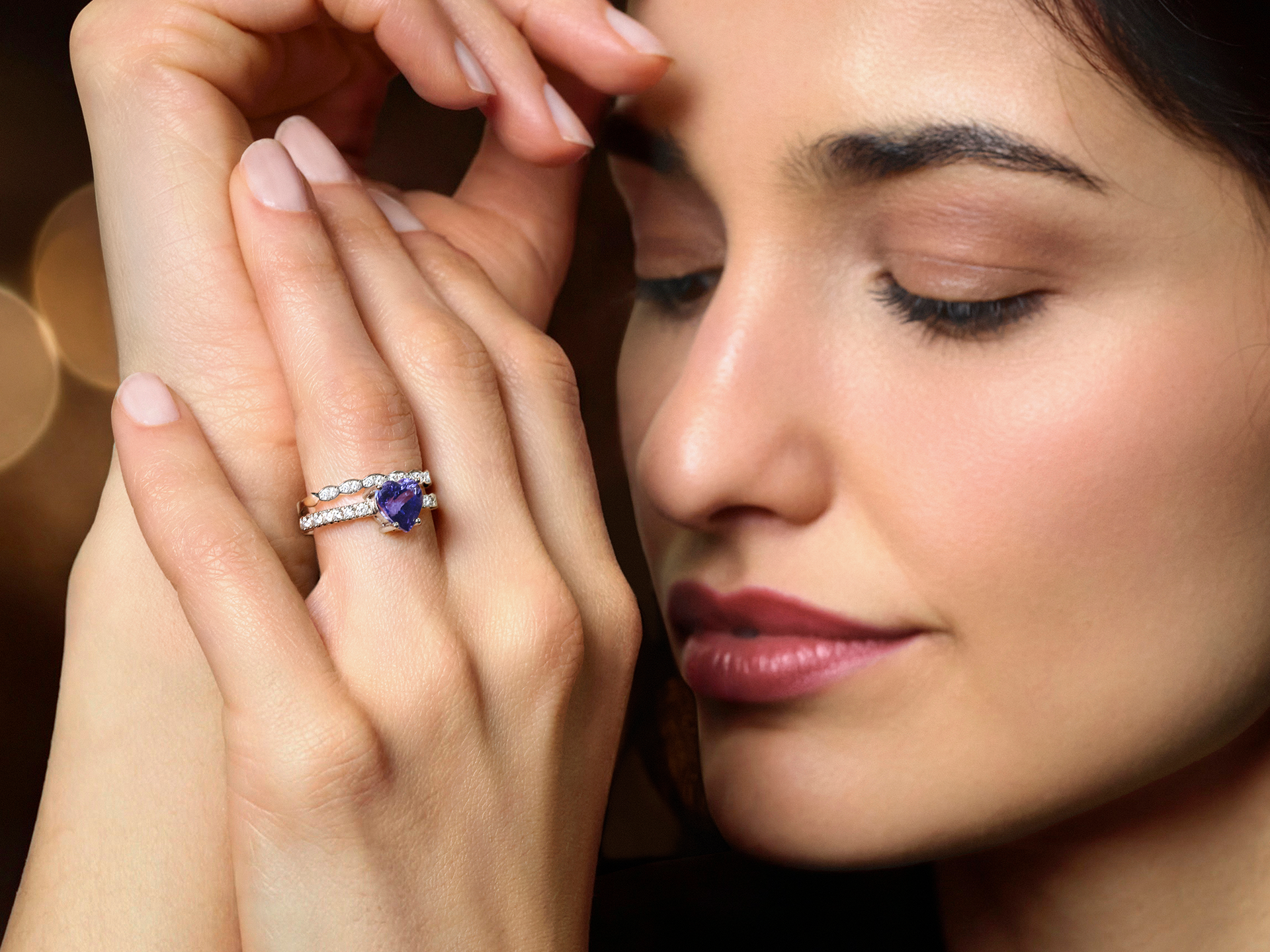 Eine Frau mit geschlossenen Augen berührt sanft ihr Gesicht und zeigt den Schullin TANSANIT RING HEART - einen herzförmigen blauen Edelsteinring mit einem Diamantring - auf ihrem Finger vor einem sanft verschwommenen Hintergrund.