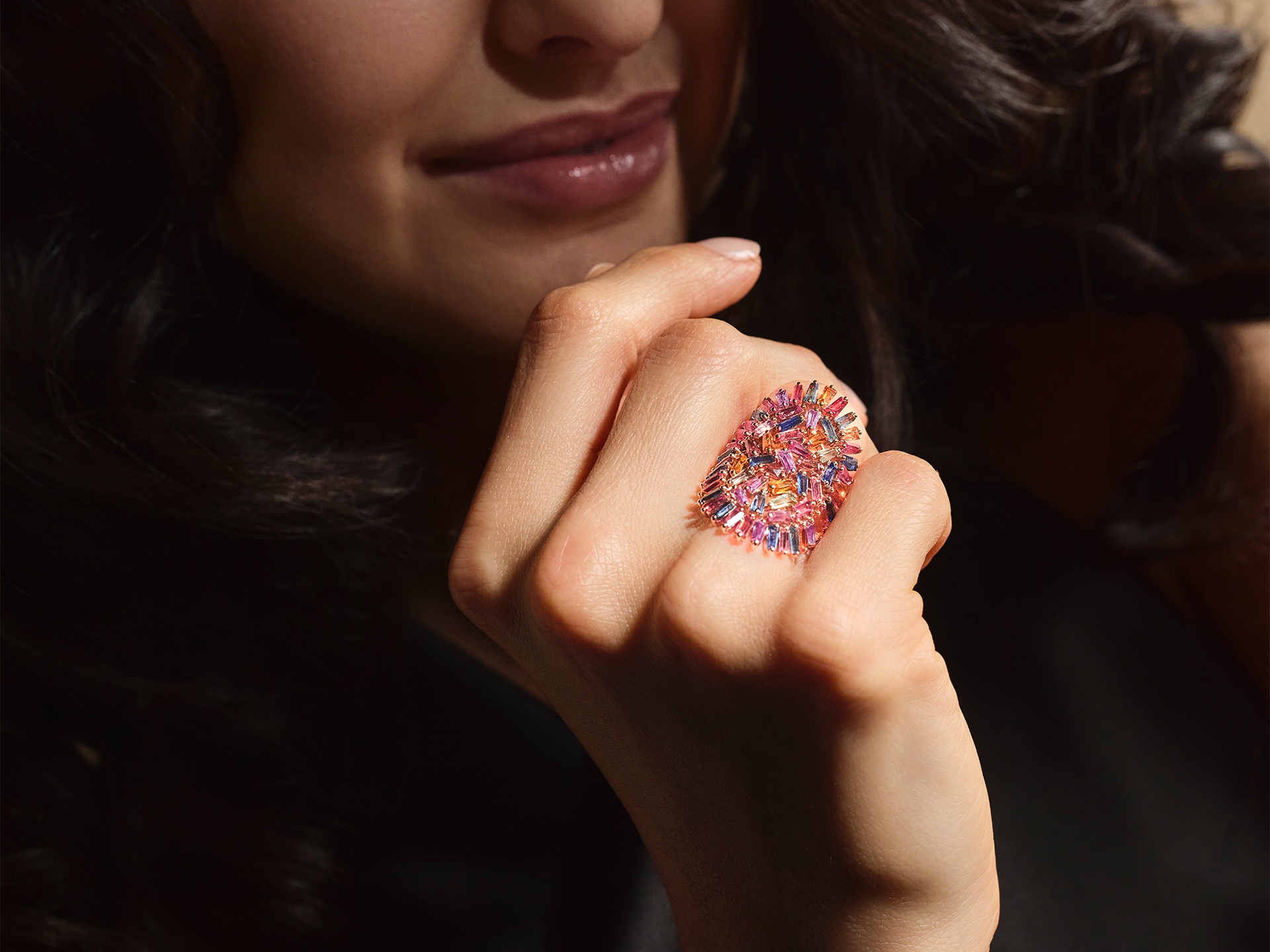 Eine Frau mit gewelltem dunklem Haar lächelt sanft und präsentiert vor einem dunklen Hintergrund den kunstvollen, mit bunten Edelsteinen besetzten RING MOSAIK von Schullin an ihrem Finger.