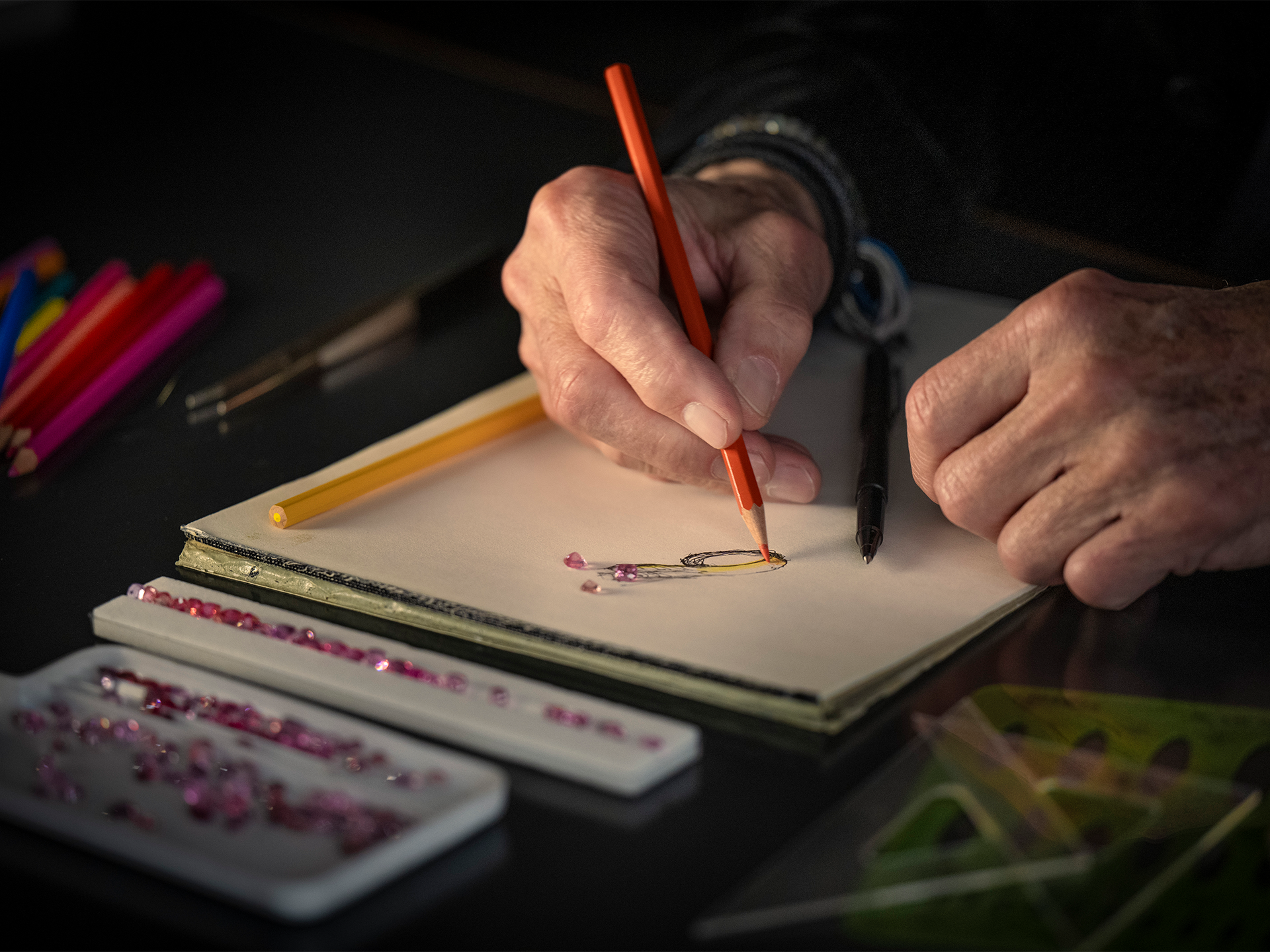 Eine Person entwirft den RING TOGETHER-Schmuck von Schullin, indem sie mit Buntstiften, roten Edelsteinen, einem Stift und Zeichenwerkzeugen auf einem dunklen Tisch skizziert.