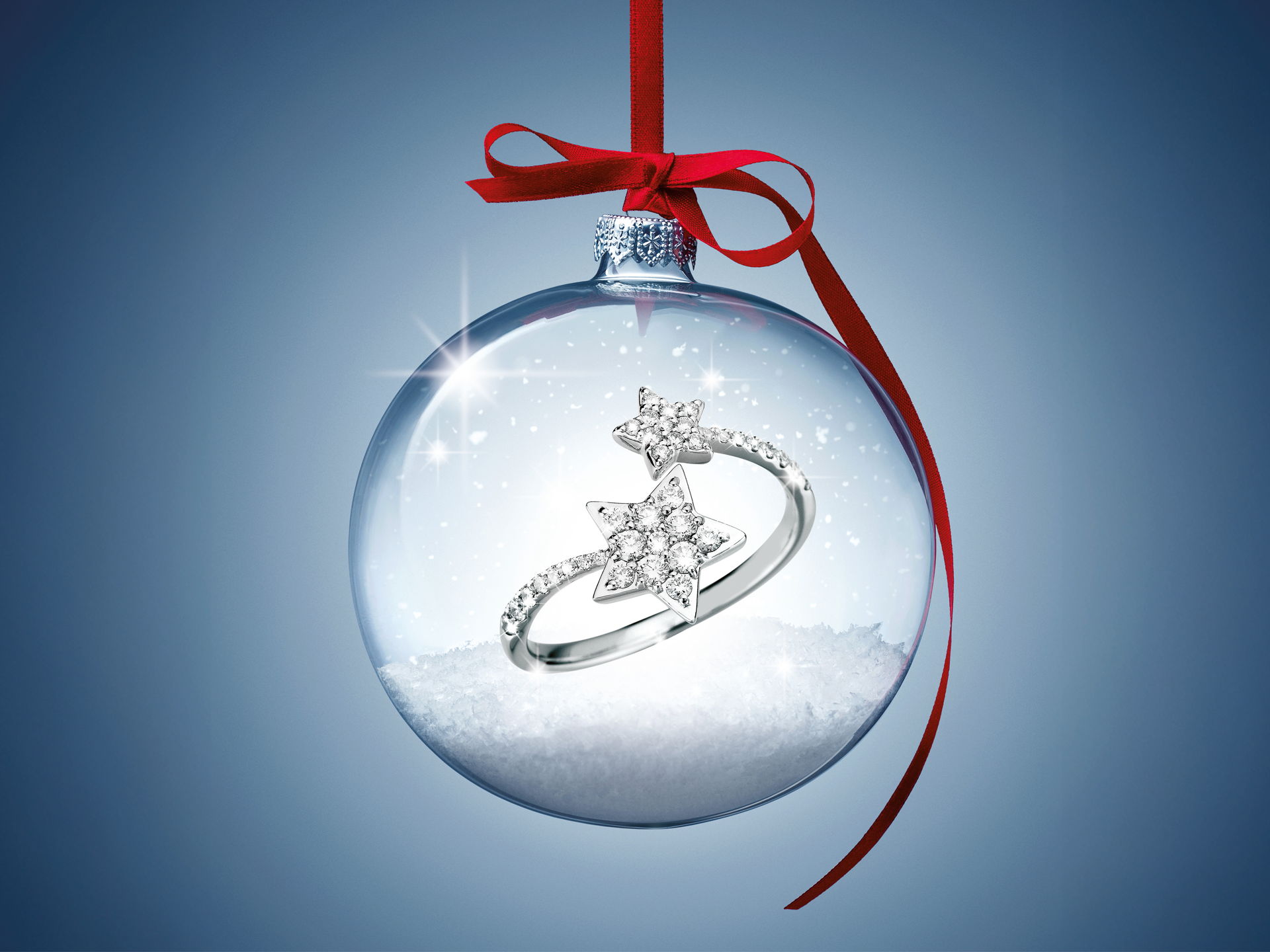 Der WEIHNACHTSSPECIAL STERNENRING von Schullin befindet sich in einem klaren Glasornament mit weißem Schnee, das an einem roten Band auf blauem Hintergrund hängt. Zwei diamantene Sternringe sind sichtbar, das Licht funkelt auf dem Glas.