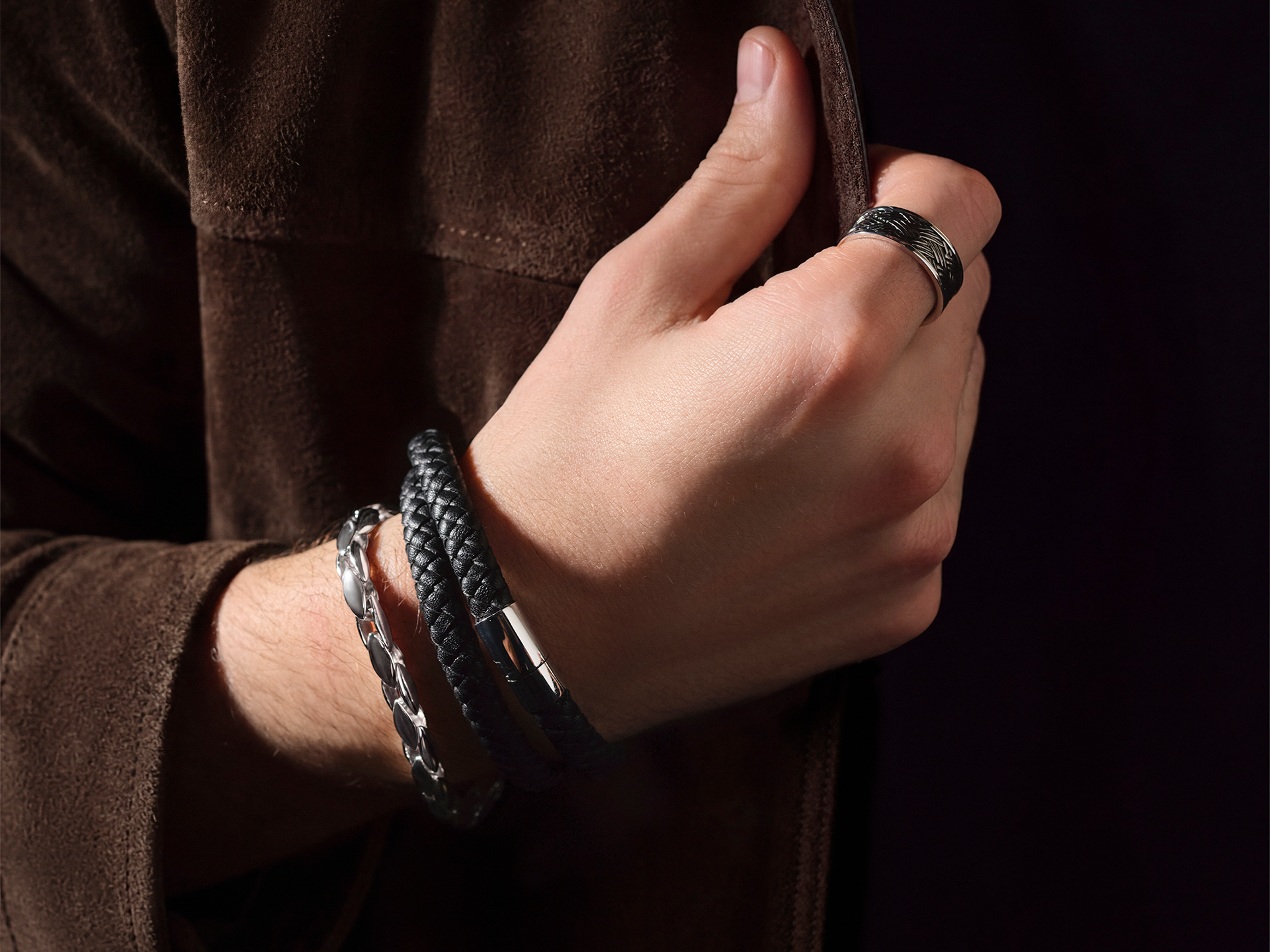 Nahaufnahme einer Hand, die das Schullin SILBER ARMBAND zusammen mit einem strukturierten Silberring und schwarzen Armbändern trägt und das Revers einer braunen Wildlederjacke vor einem dunklen Hintergrund umgreift.