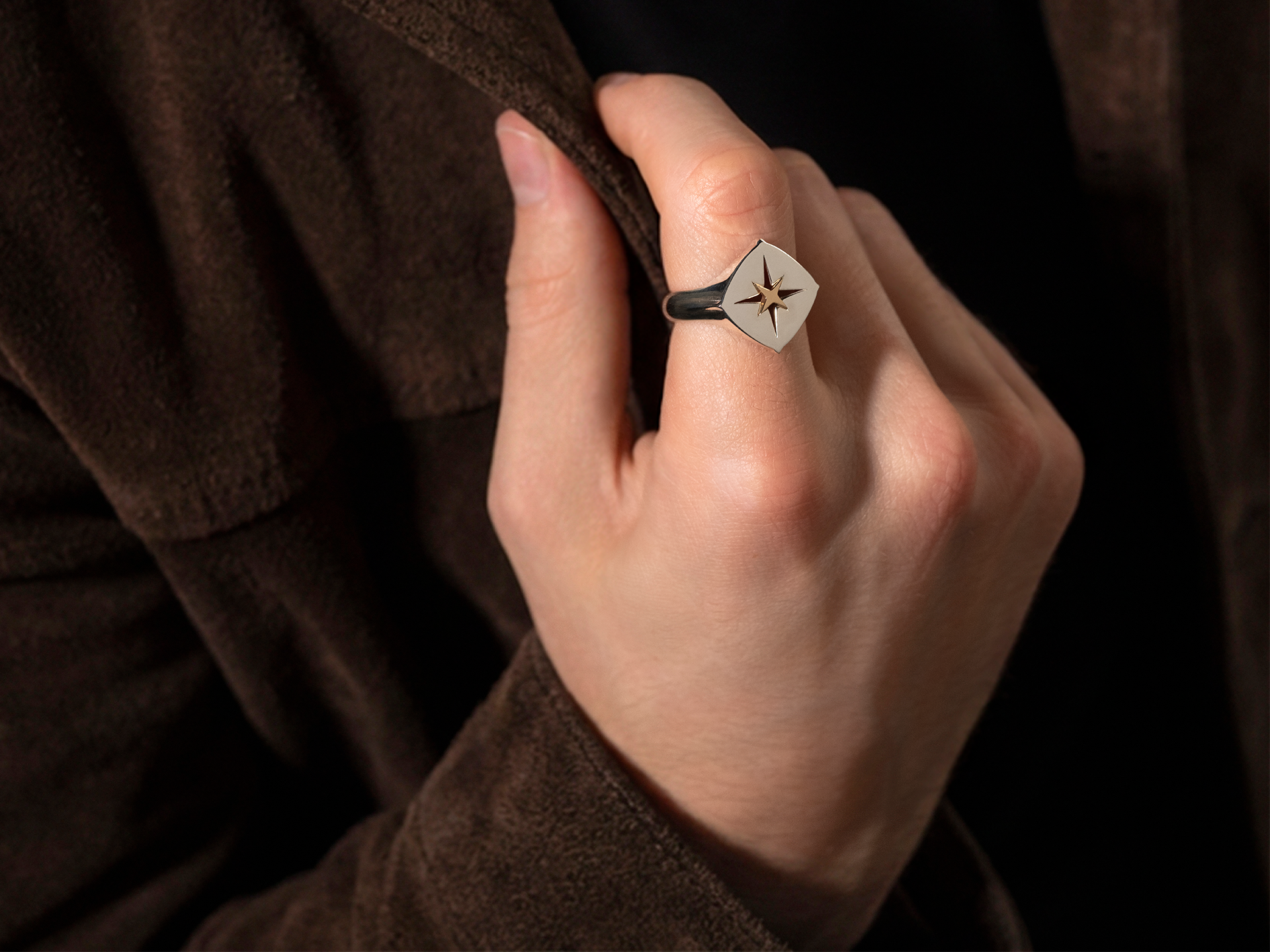 Nahaufnahme einer Hand, die den Schullin ELEMENTS RING SILBER trägt, einen quadratischen Silberring mit schwarzem Sternmuster, der auf einer braunen Wildlederjacke abgebildet ist.