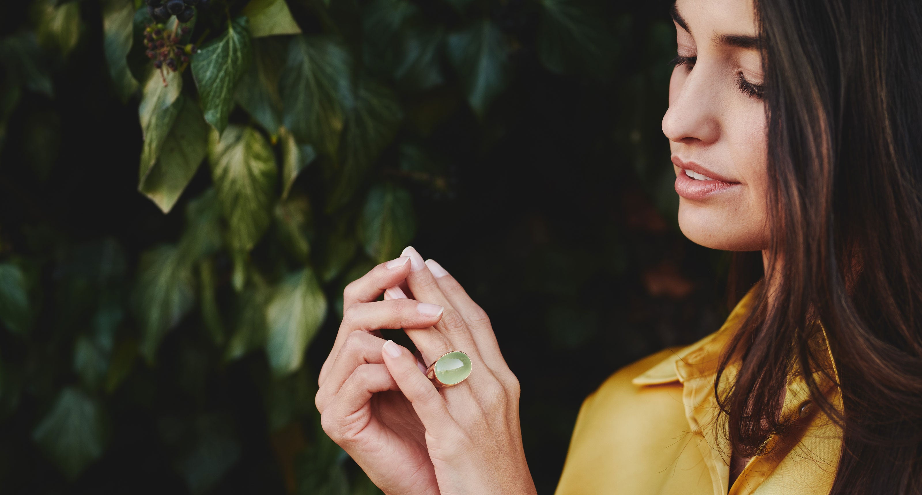Eine Frau in einem gelben Hemd schaut auf einen großen grünen Ring an ihrem Finger und steht vor dunkelgrünen Blättern.